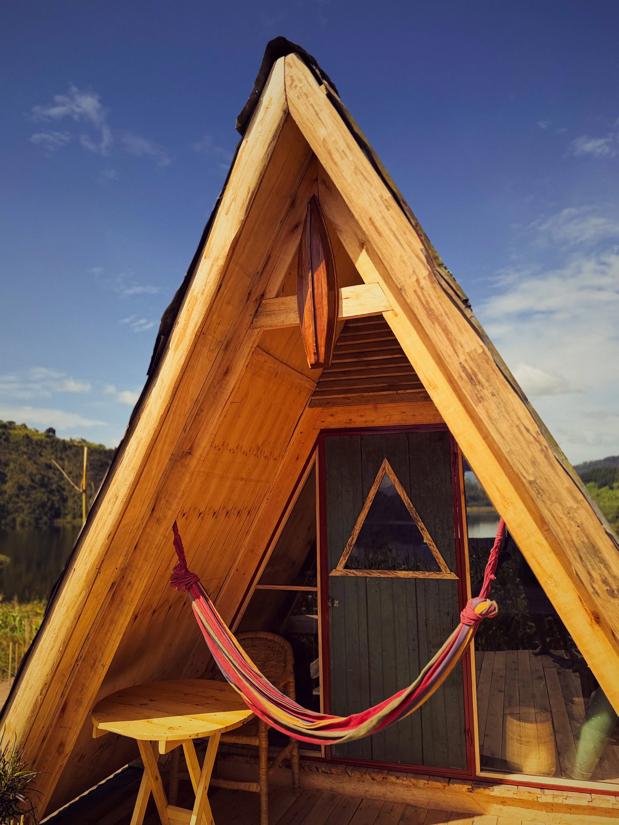 A small, wooden A-frame cabin with a hammock, table, and chair outside, near a lake and green hills under a blue sky.