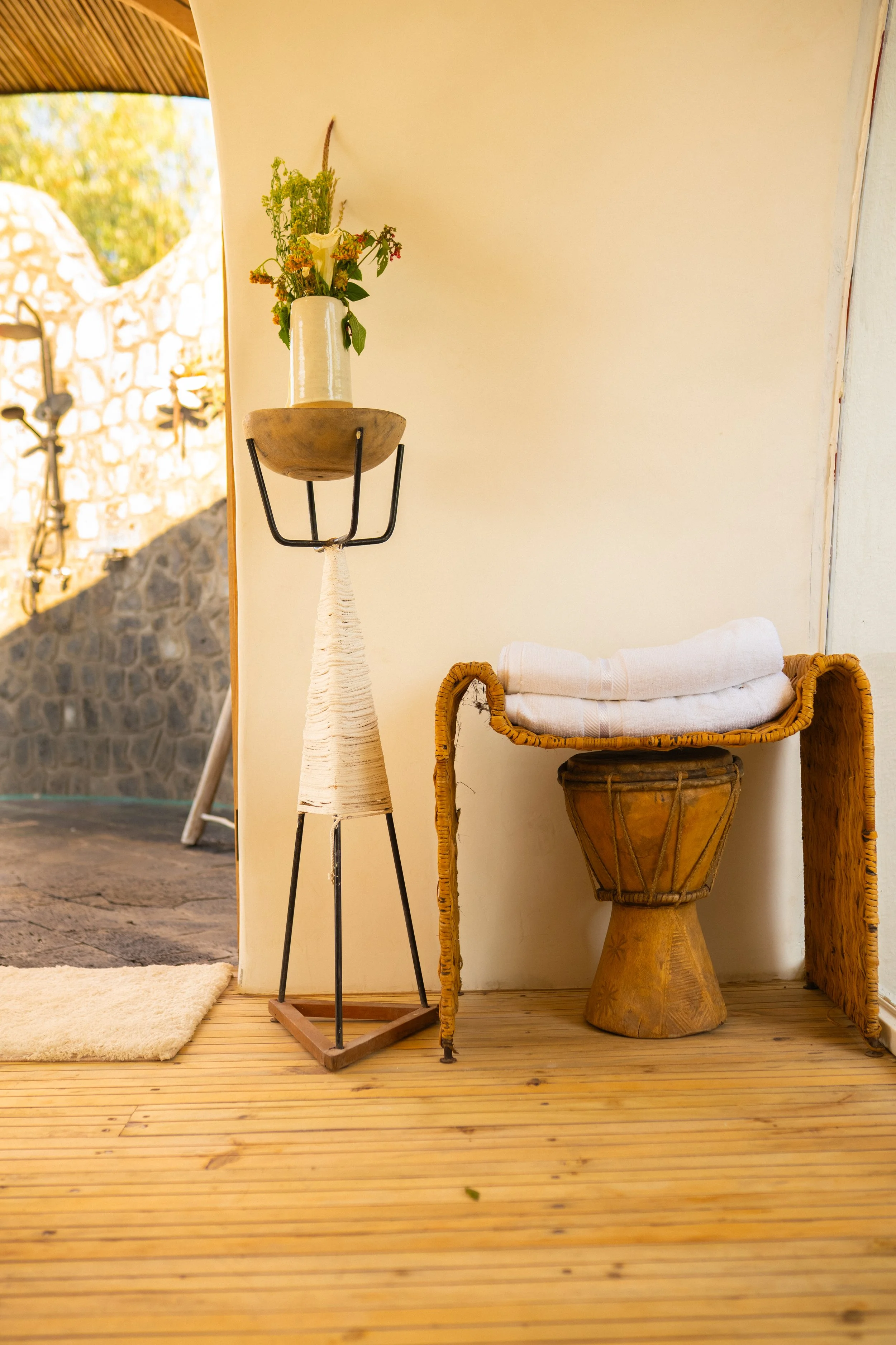 A corner with wooden flooring, featuring a small beige rug, a tall floor lamp with a bouquet of flowers in a white vase, a traditional drum with white cloth on top, and a woven screen.