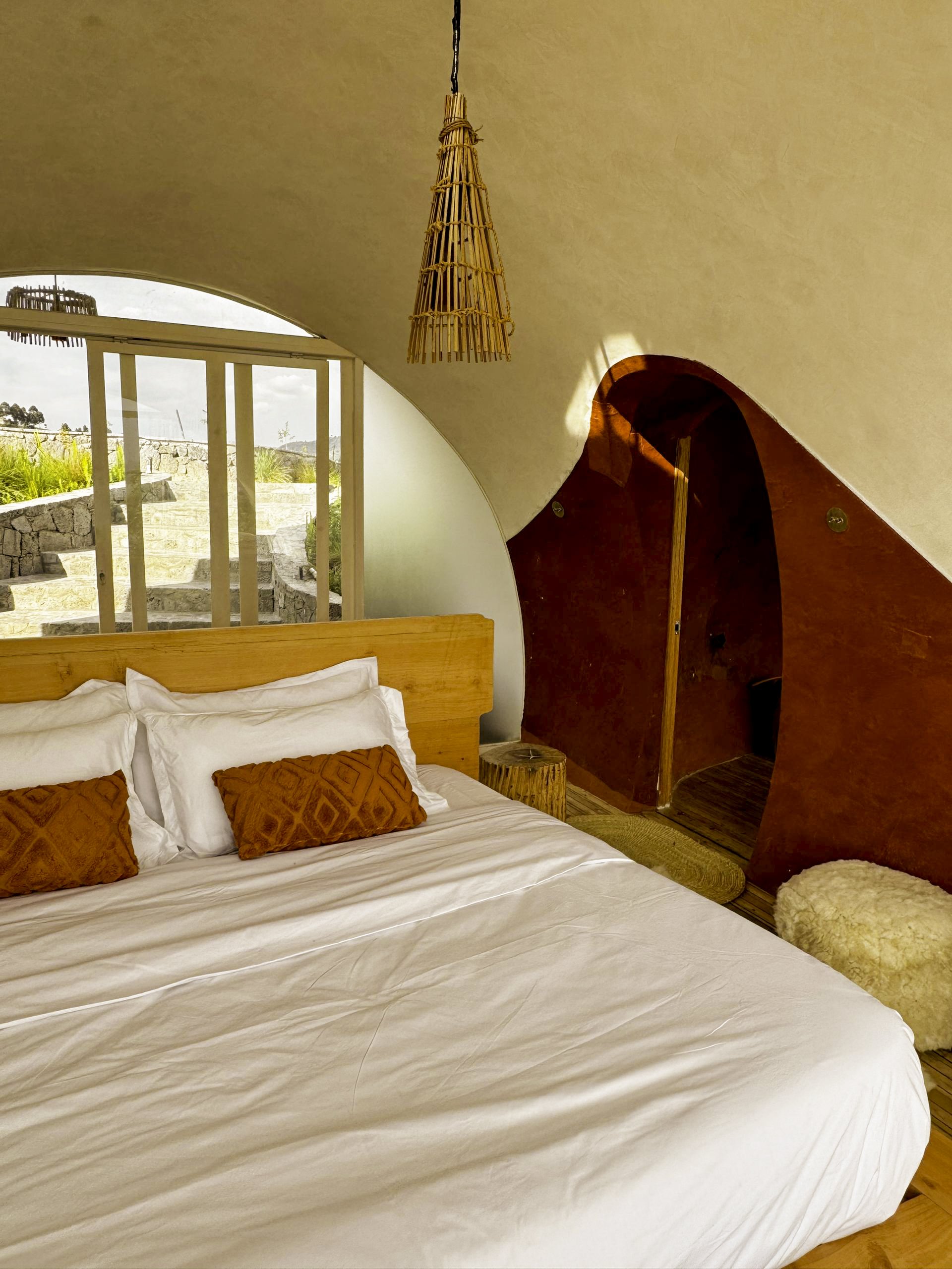A cozy bedroom with a wooden bed, white linens, and orange decorative pillows. There is a window with a view of stone stairs outside, and a small round wooden side table. The walls are painted red and beige, with unique curved architecture. A woven hanging lamp is suspended from the ceiling.