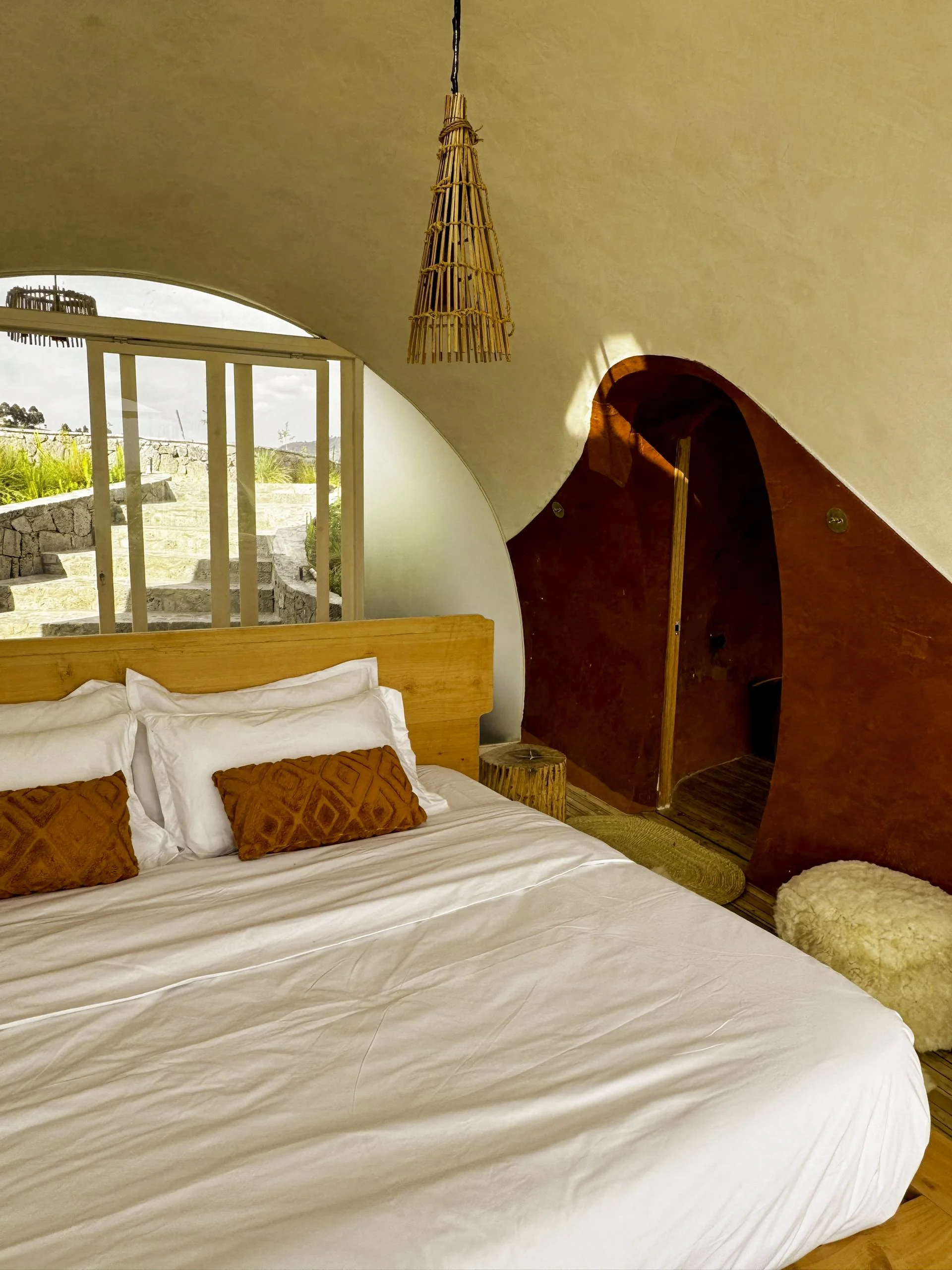 A cozy bedroom with a large bed featuring white linens and two orange decorative pillows. There is a wooden headboard and a small wooden side table. The room has a curved ceiling with a woven pendant light hanging from it. An arched doorway leads to 