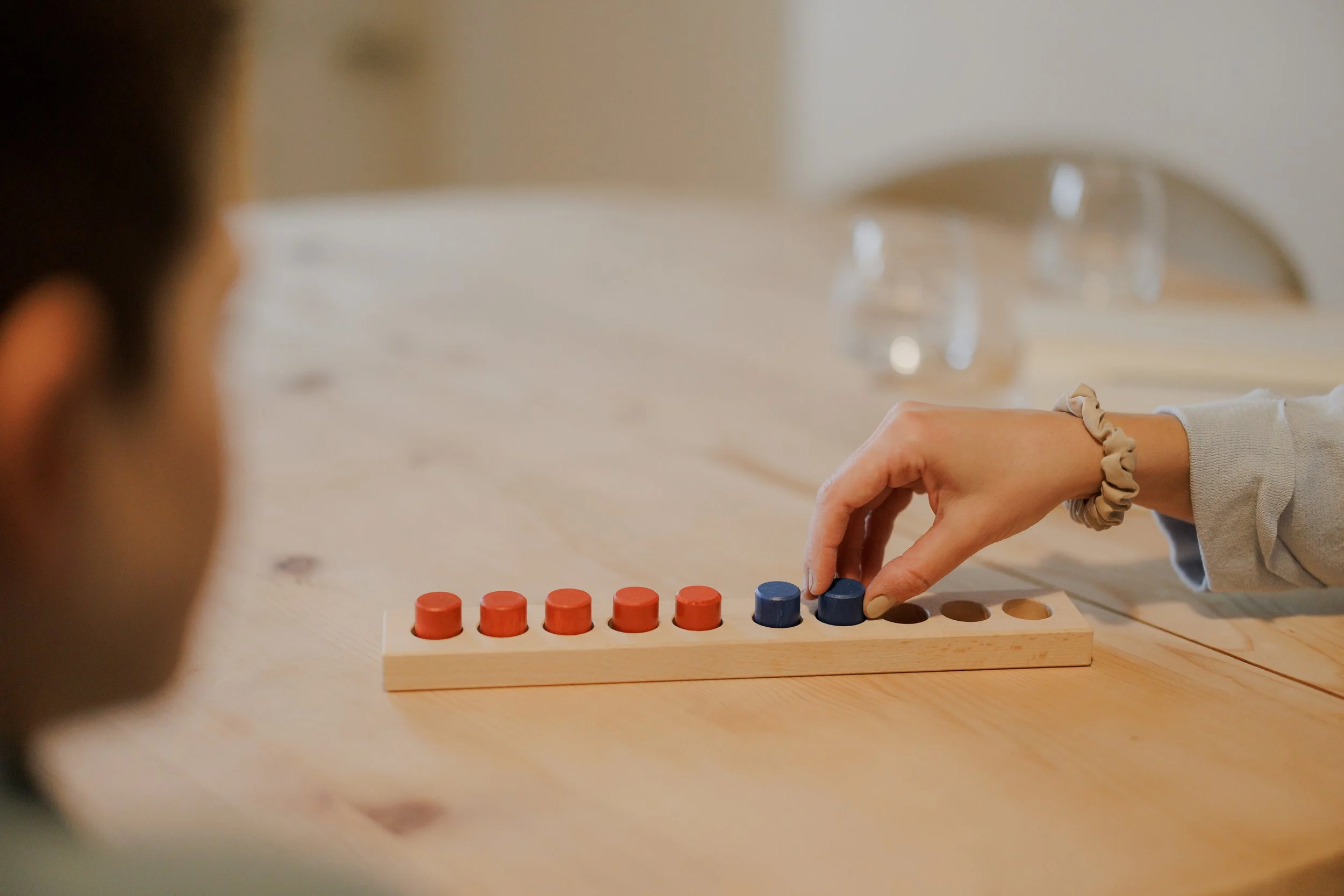 Nahaufnahme einer Person, die ein Spiel mit bunten Holzklötzen auf einem Holztisch spielt, während eine andere Person im Vordergrund beobachtet.