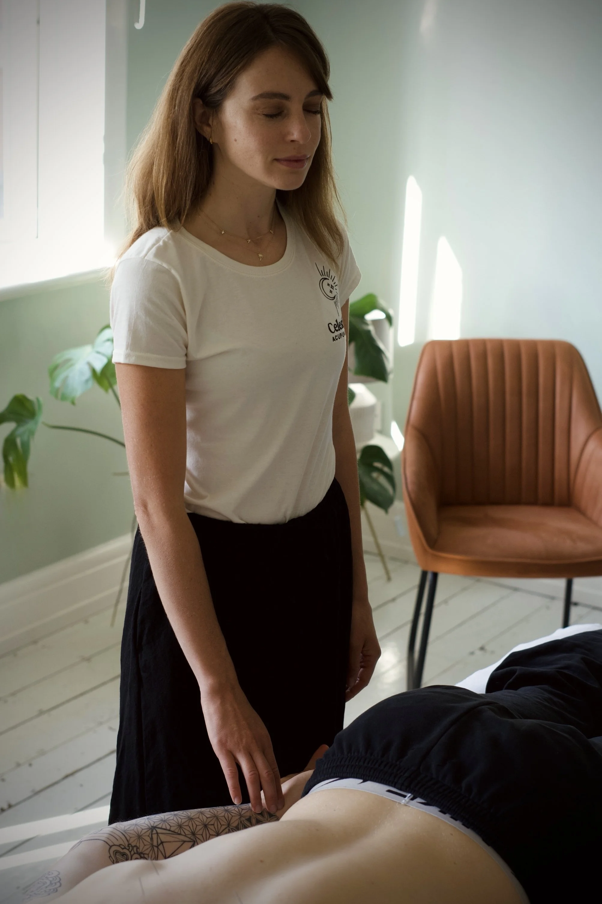 A woman standing next to a bed, looking down at a person lying on the bed in a massage or therapeutic setting with plants and a brown chair in the background.