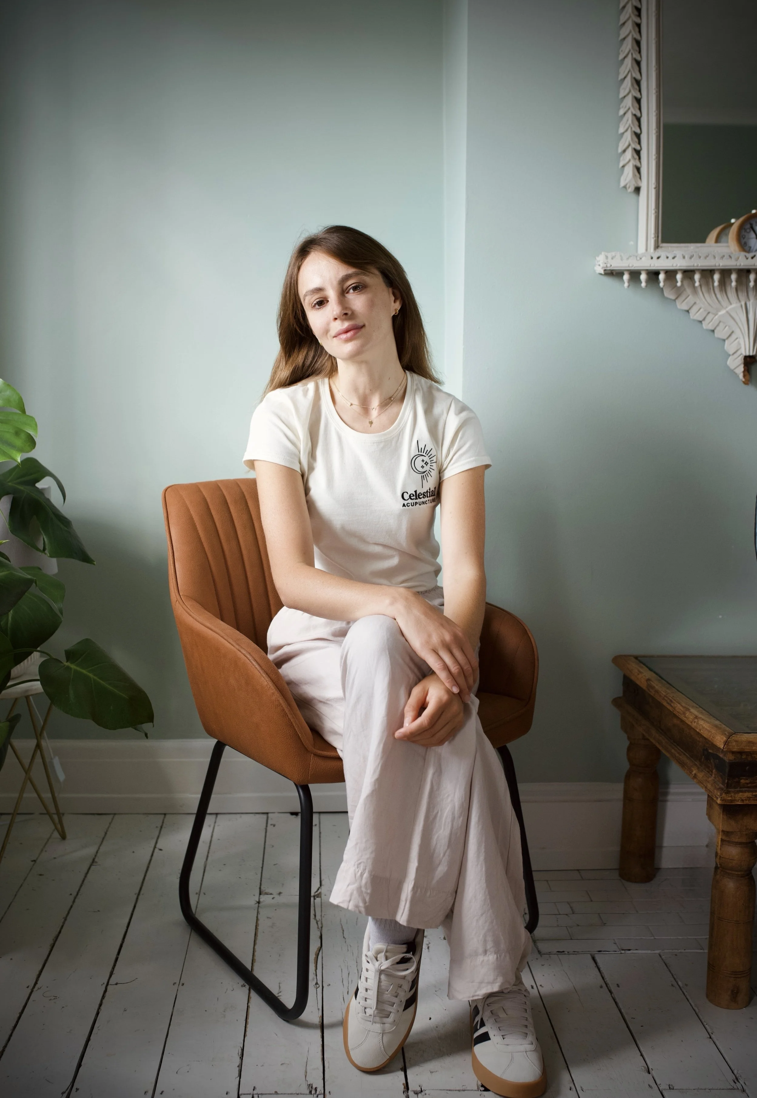 A young woman sitting on a tan upholstered chair with black metal legs in a room with light green walls, a white wooden floor, and a rustic wooden table. She wears a white t-shirt with a small black and white logo on the chest, off-white wide-leg pants, and white sneakers. She has long brown hair and a neutral expression.