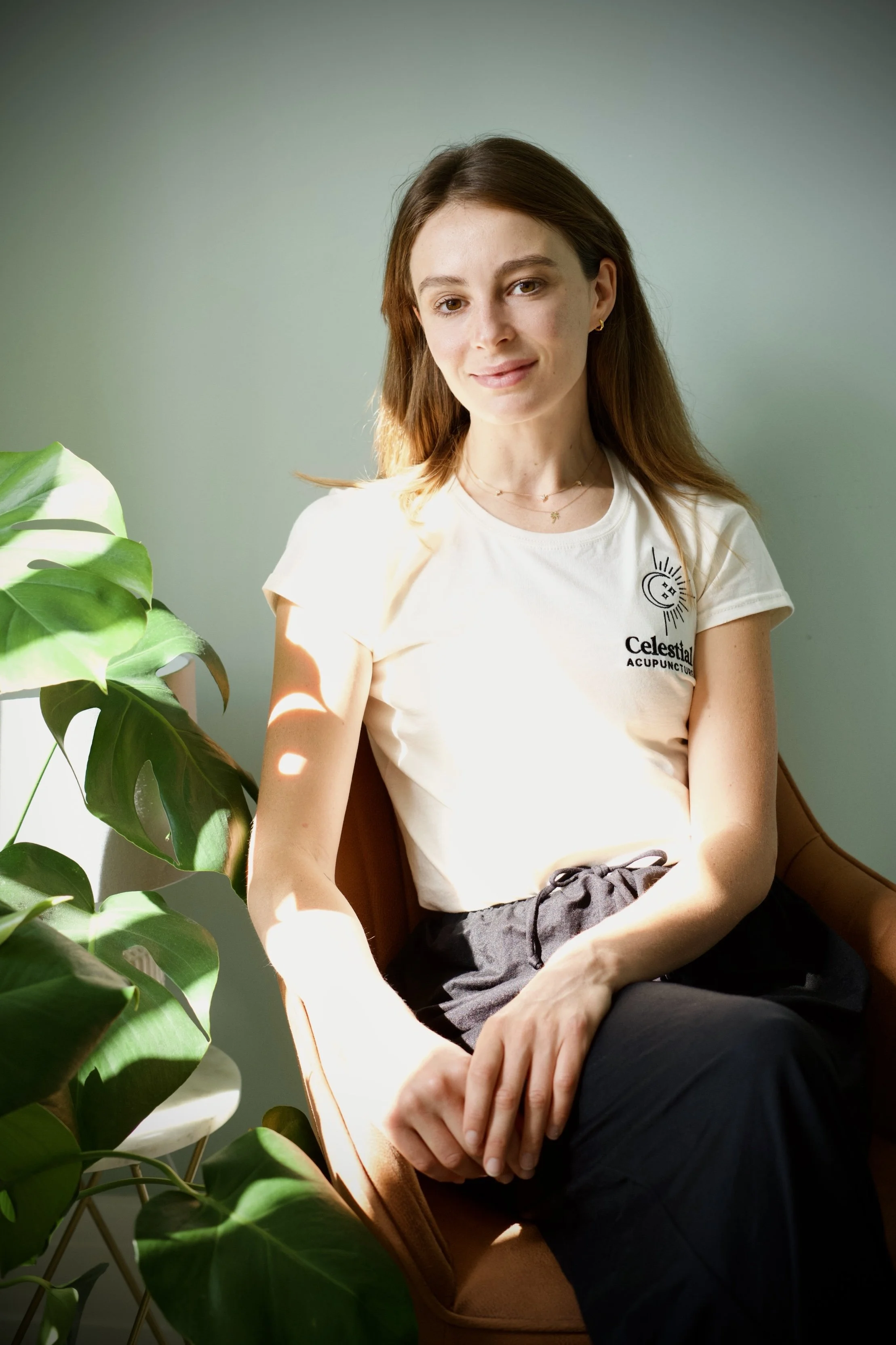 A woman sitting in a chair next to a green plant, wearing a white t-shirt with a logo that says 'Celestial Acupuncture,' and black pants, in a softly lit room.