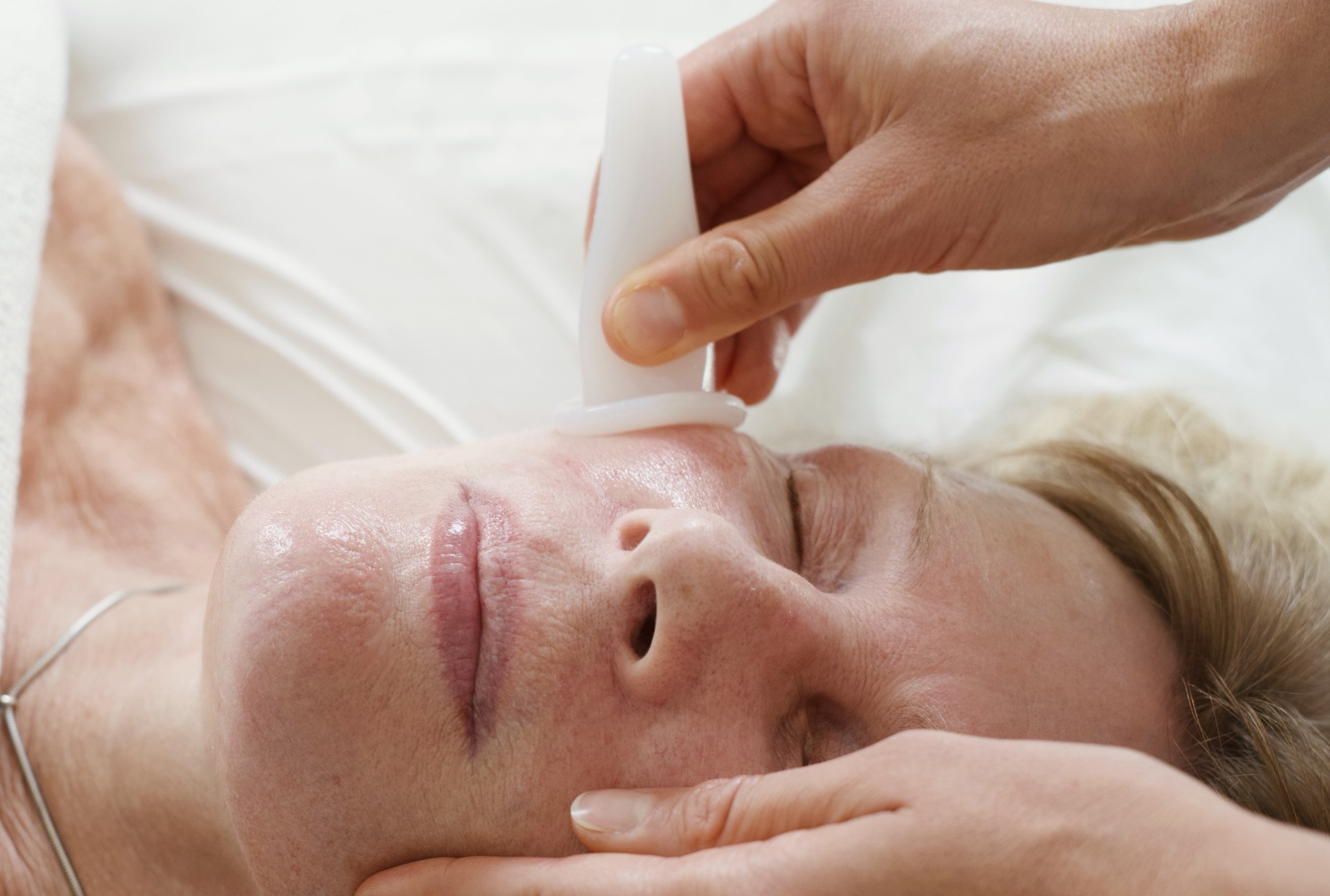 A woman lying with eyes closed receiving a facial treatment from a professional using a white spatula or roller on her face.