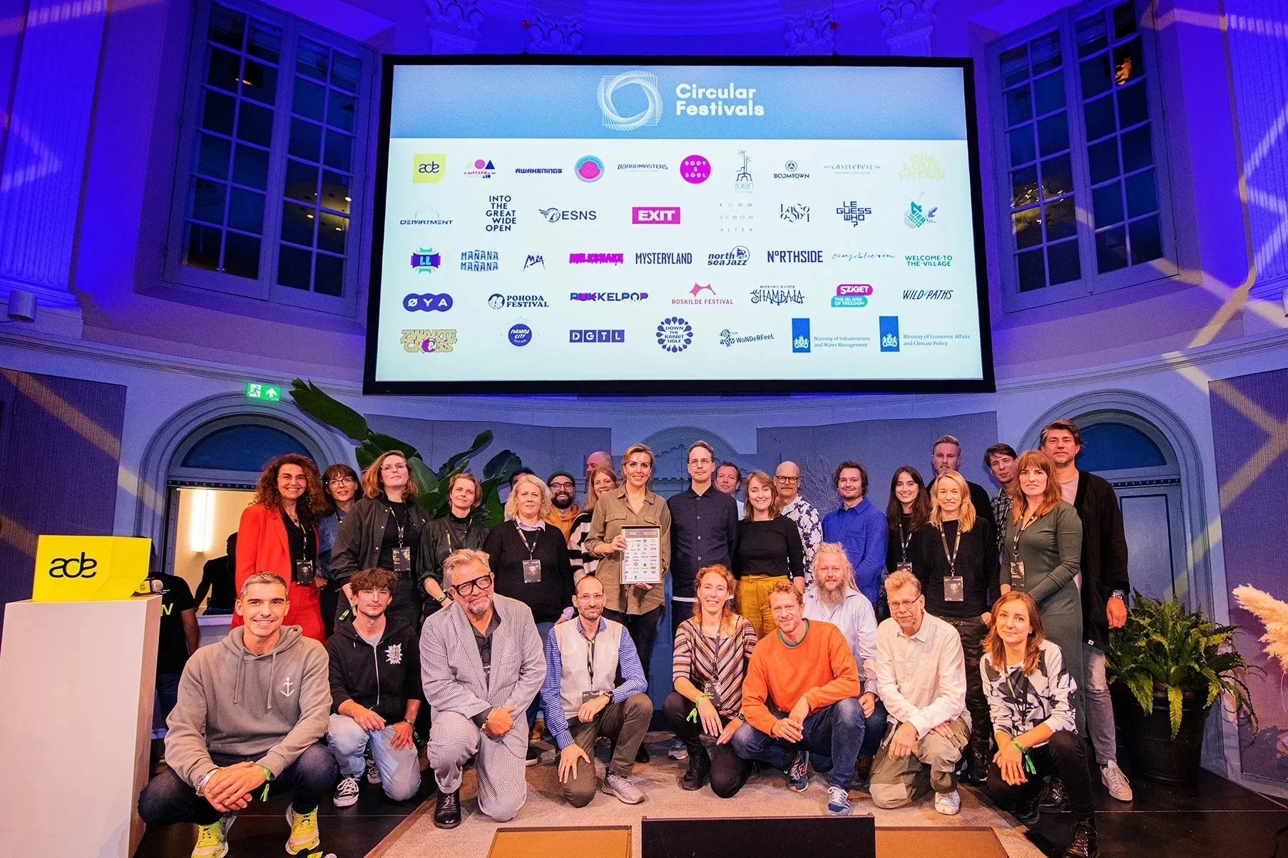 Groep mensen poseert voor een scherm met logo's van festivals, in een grote zaal met hoge ramen en blauwe verlichting.