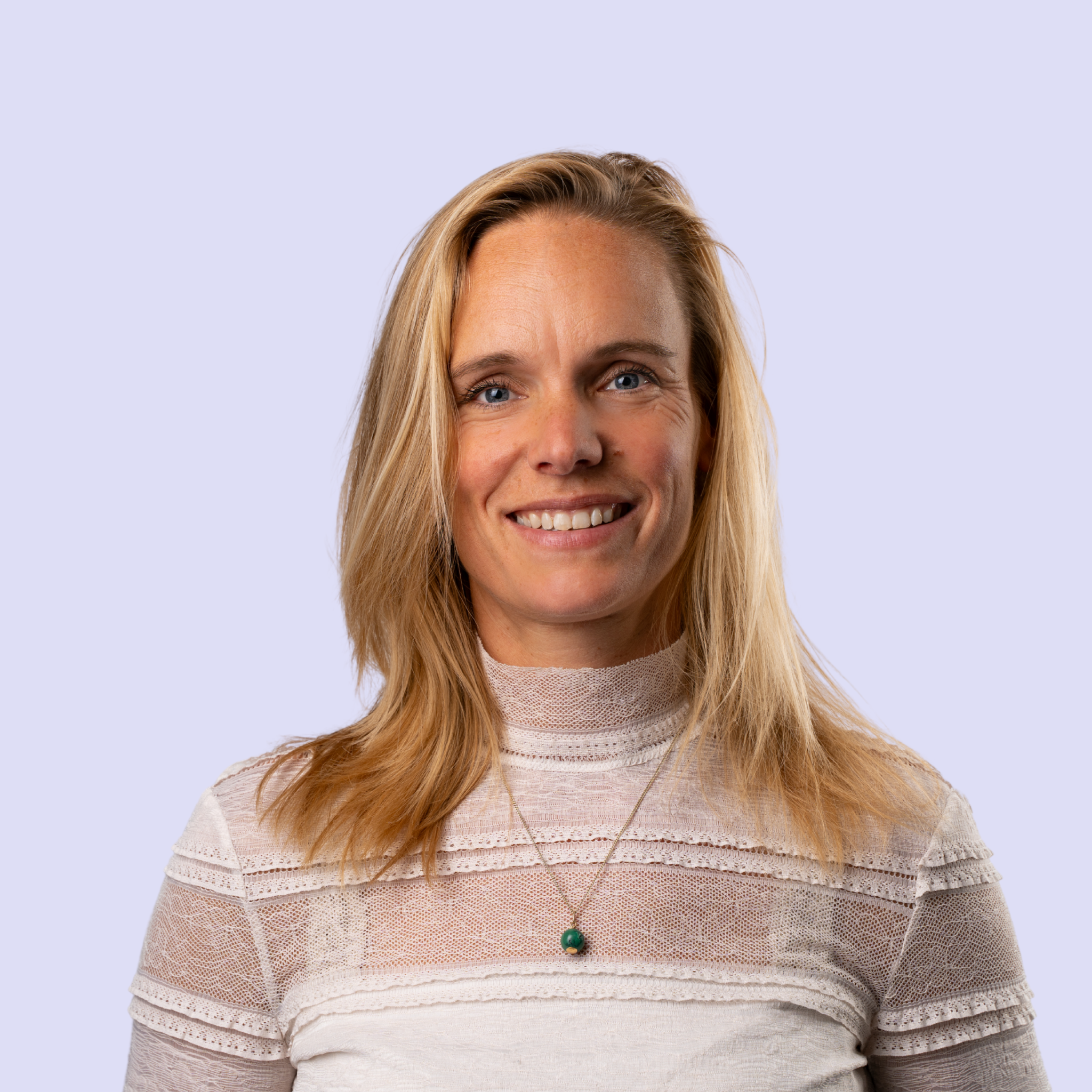 Portret van een Lyke Poortvliet, lachende blonde vrouw met blauwe ogen, zittend voor een lichtpaarse achtergrond.