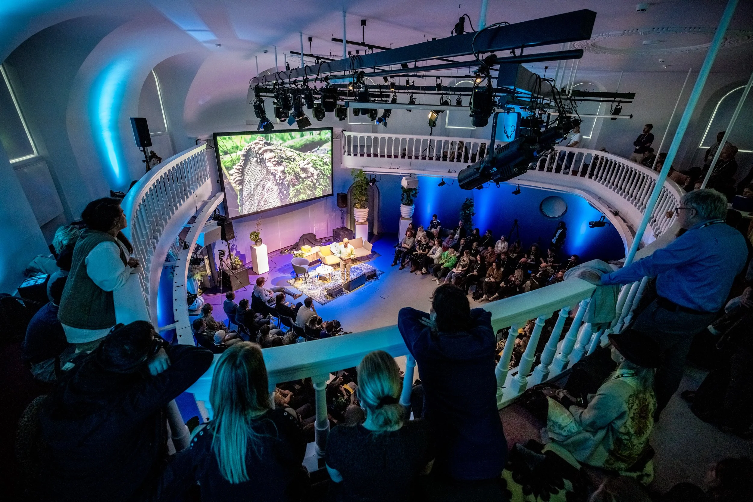 Binnenoptreden in een auditorium met een publiek, een houten podium met stoelen en een grote scherm dat een buitenlandschap toont, met ronde balkonranden en professionele belichting.