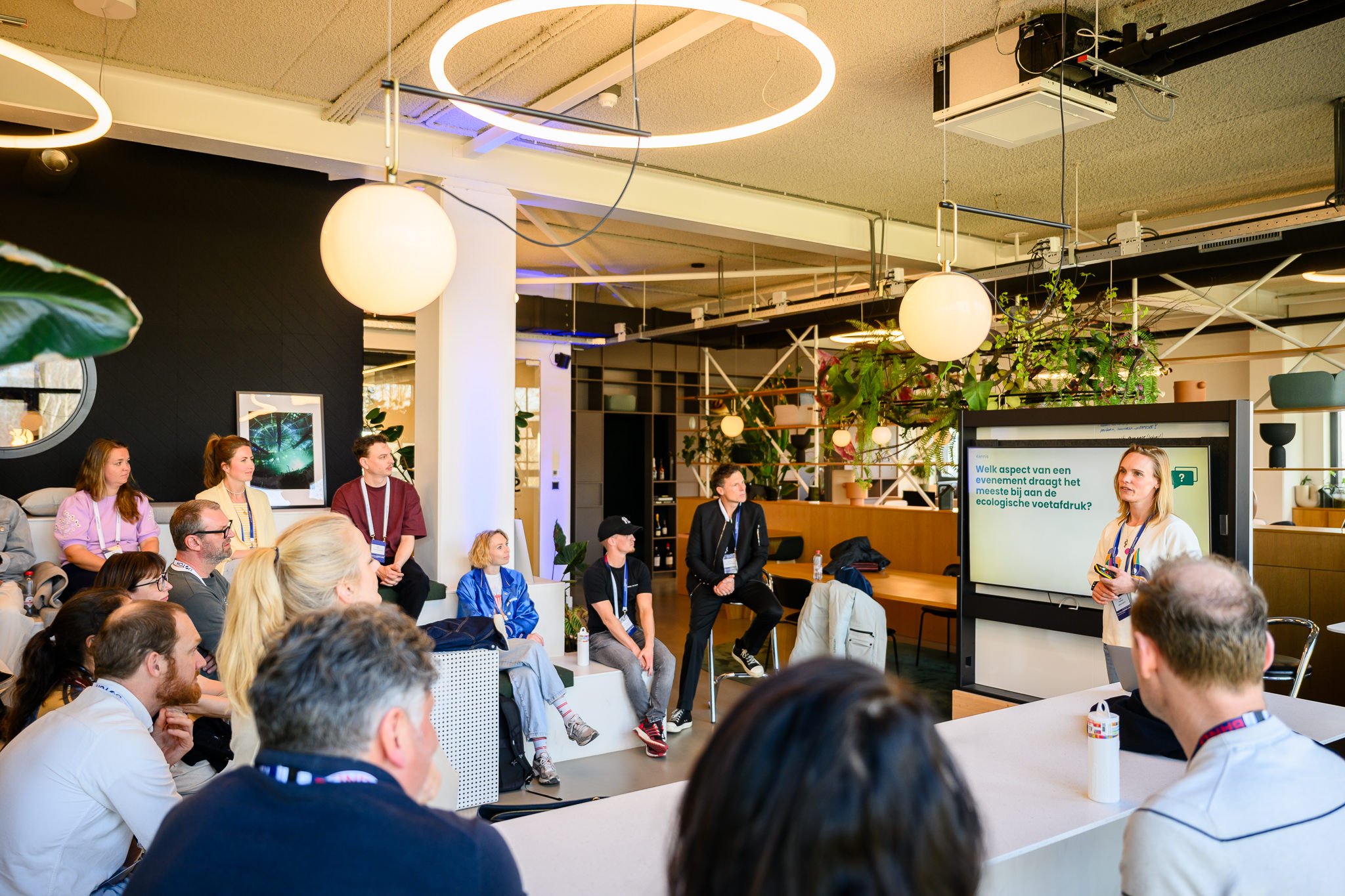 Een vrouw geeft een presentatie aan een groep mensen in een moderne, groene vergaderruimte. Op de schermen staan vragen over ecologische voetafdruk.