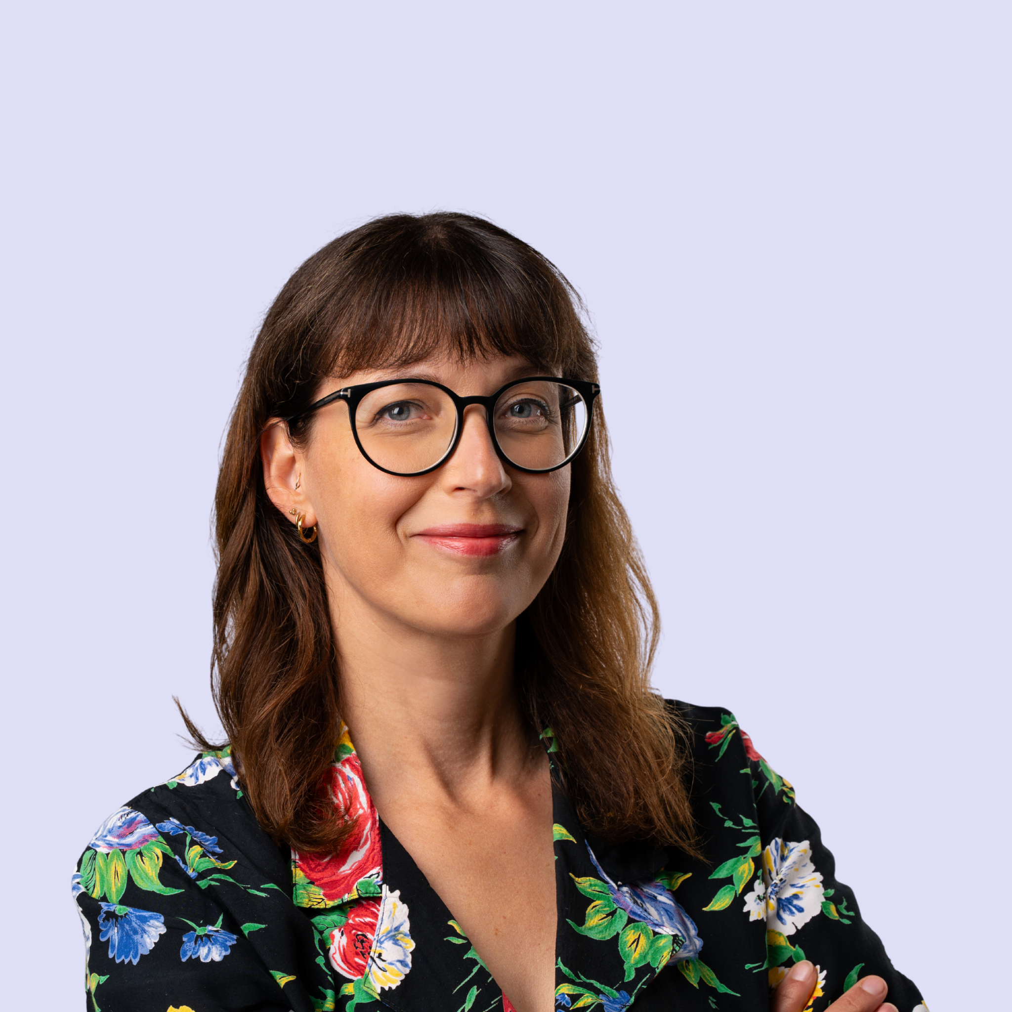 Een portretfoto Laura van de Voort,  een vrouw met bruin haar, zwart-rond bril en een zwarte blouse met een kleurrijk bloemenpatroon, lachend en met de armen gekruist tegen een lichtblauwe achtergrond.