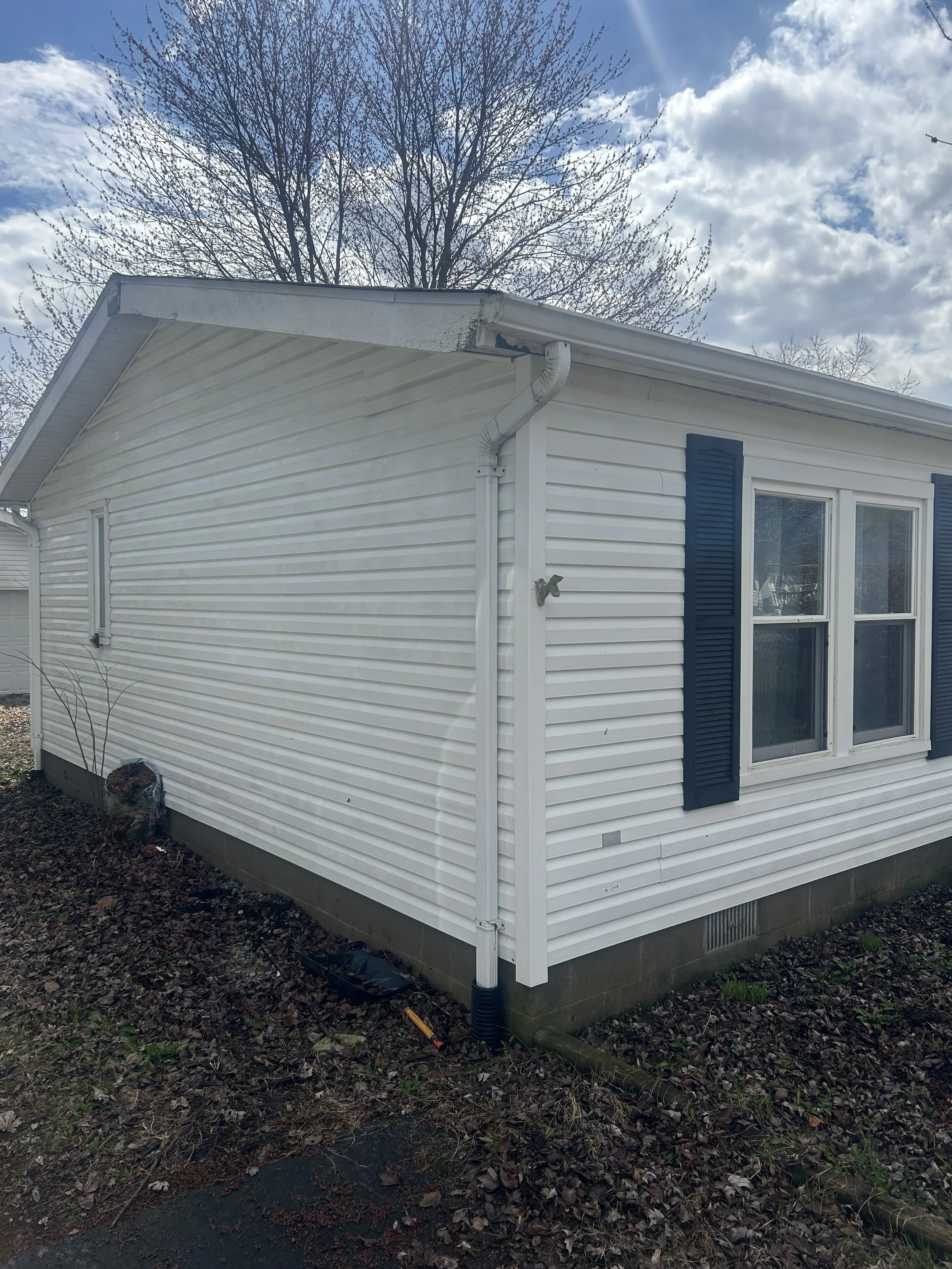 Front corner of a white vinyl-sided house with blue shutters, a window, and a leafless tree in the background; cloudy sky overhead.