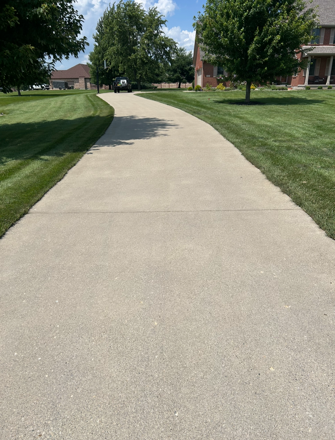 A curving concrete sidewalk through a residential yard with green grass, trees, a house, and parked vehicles in the background.