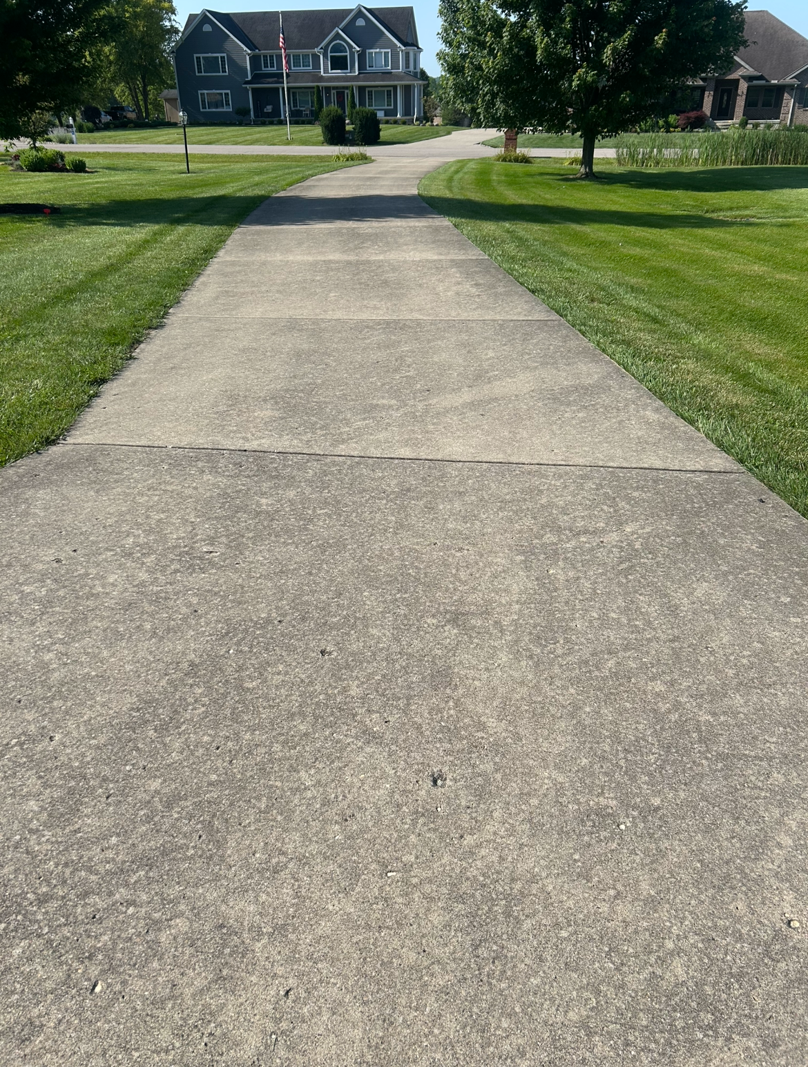 Concrete sidewalk leading to a residential area with houses, trees, and well-maintained lawns on a sunny day.