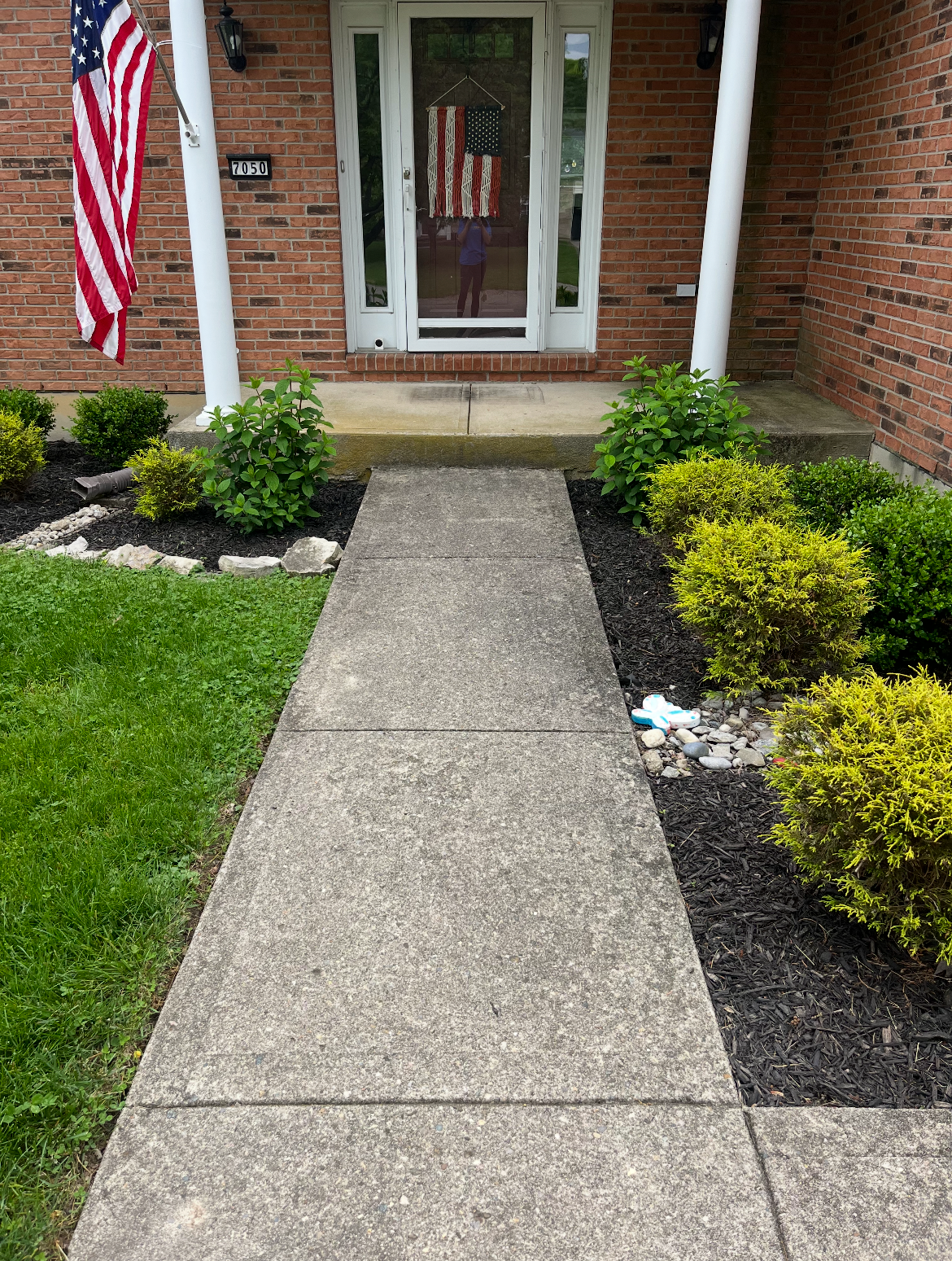 Concrete pathway leading to a front door with a glass screen door. Garden beds with bushes and rocks on either side of the pathway. An American flag hanging on a pole to the left.