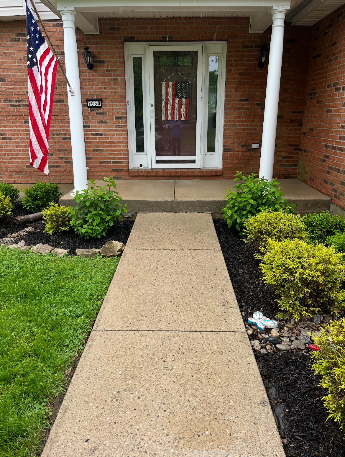 Front porch of a brick house with a concrete walkway leading to a white front door, American flag hanging on the left, and a decorative American flag hanging inside the door. Green shrubbery borders the walkway on both sides.