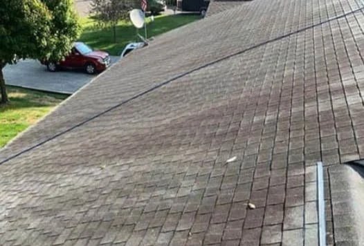 Close-up view of a brown wooden shingle roof with a small chimney in the distance under a clear blue sky.