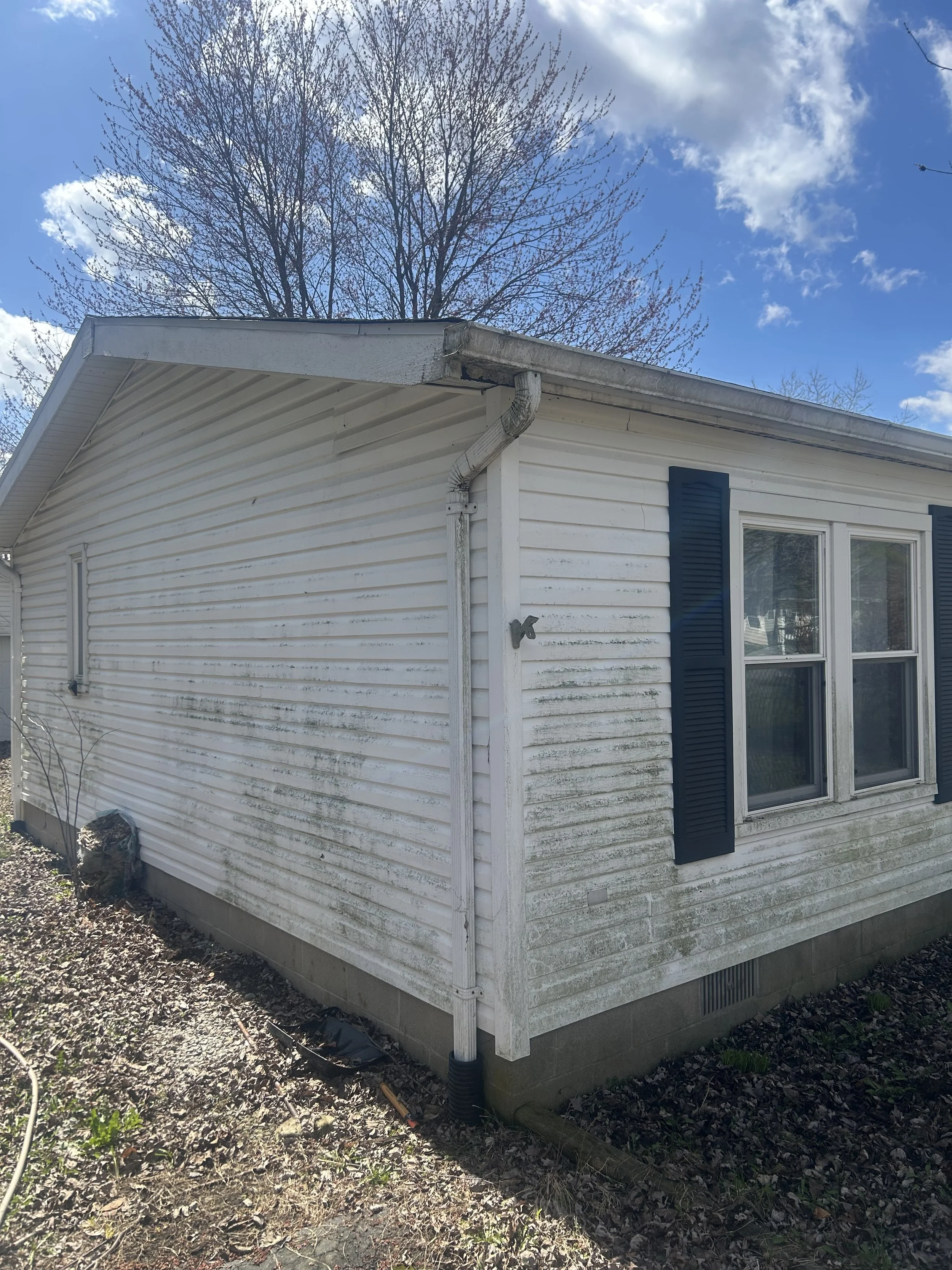 The exterior of a white house with blue shutters and dirty siding, showing a corner, a window, and a downspout under a partly cloudy sky.