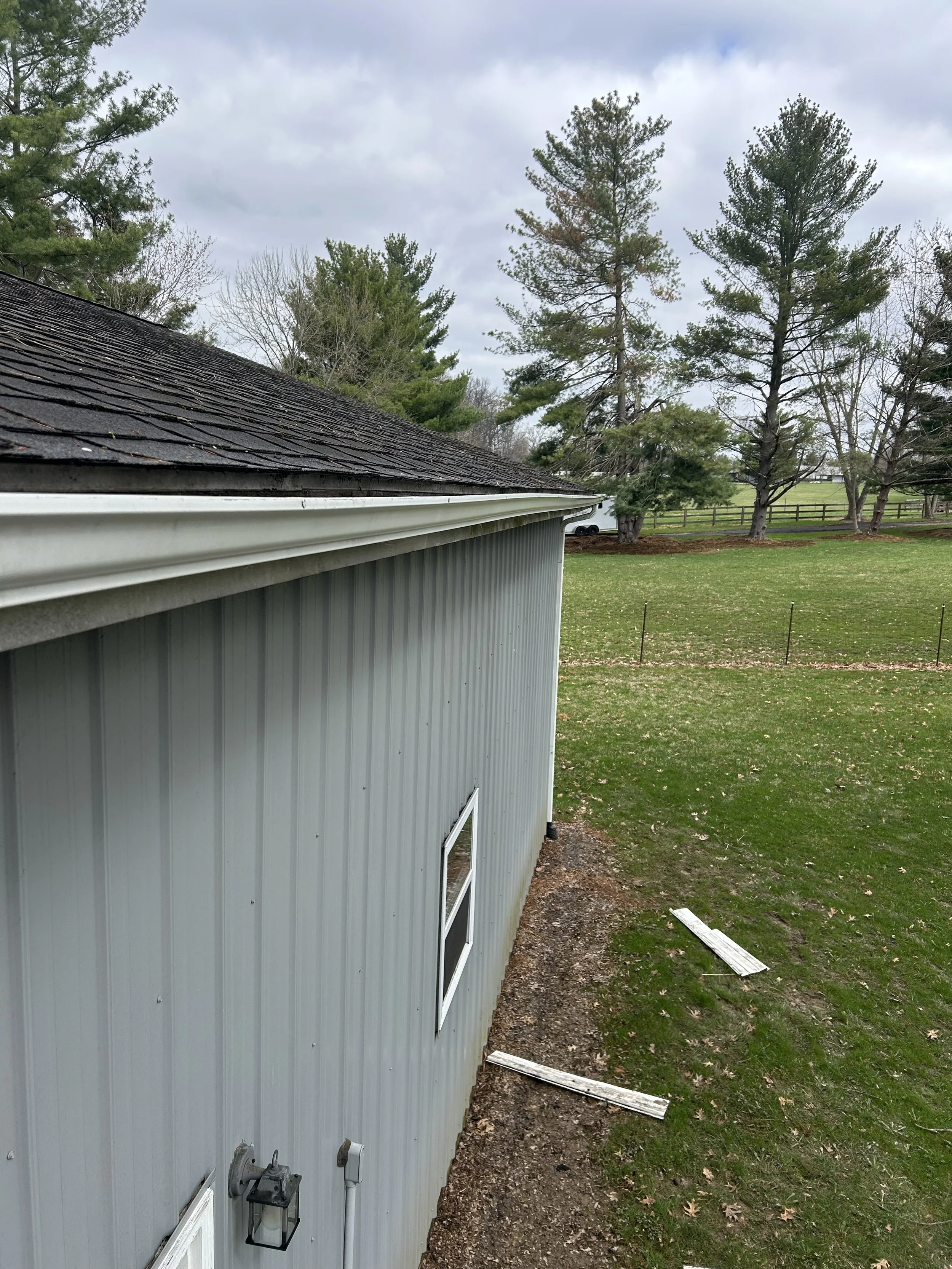 Side of a metal building with a window, gutter, and a light fixture, surrounded by a grassy yard with trees and a cloudy sky.