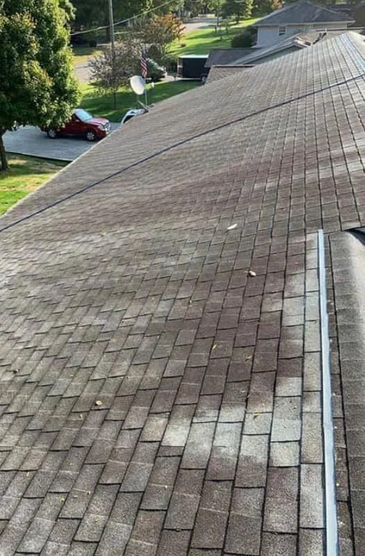 View of a sloped residential roof with brown shingles, a neighboring house, trees, parked cars, and an American flag in the background.