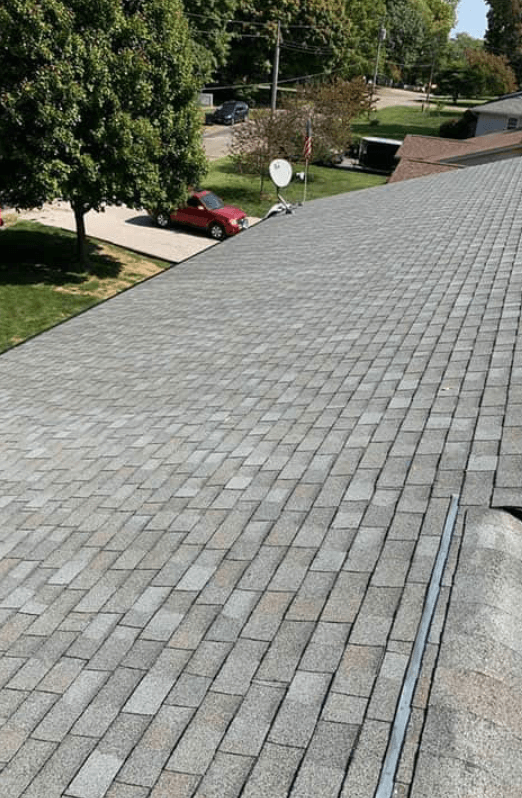 View of a roof covered with gray asphalt shingles, extending towards the horizon, with trees, a parked red car, satellite dishes, and a street with trees and utility poles in the background.