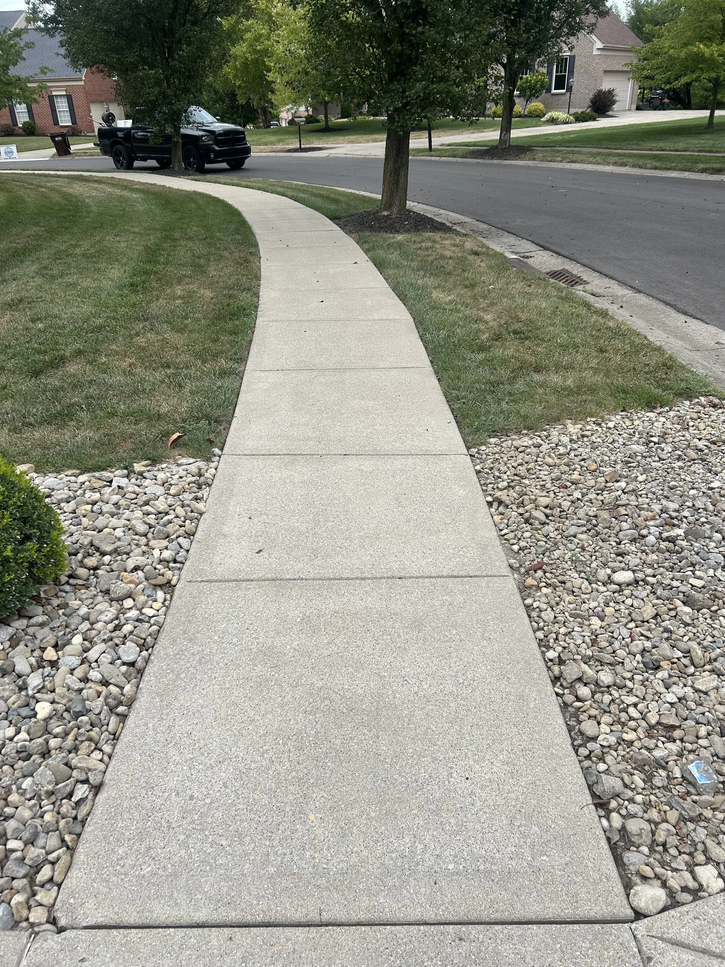 Concrete sidewalk lined with small rocks and grass, curving along a residential street with trees and houses in the background.