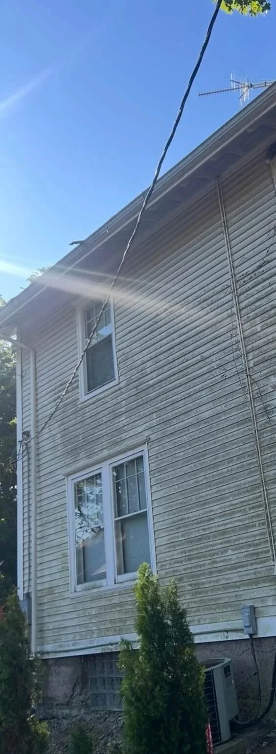Side of a house with weathered beige siding, two windows, and an air conditioning unit at the base, with a clear blue sky and laundry line.