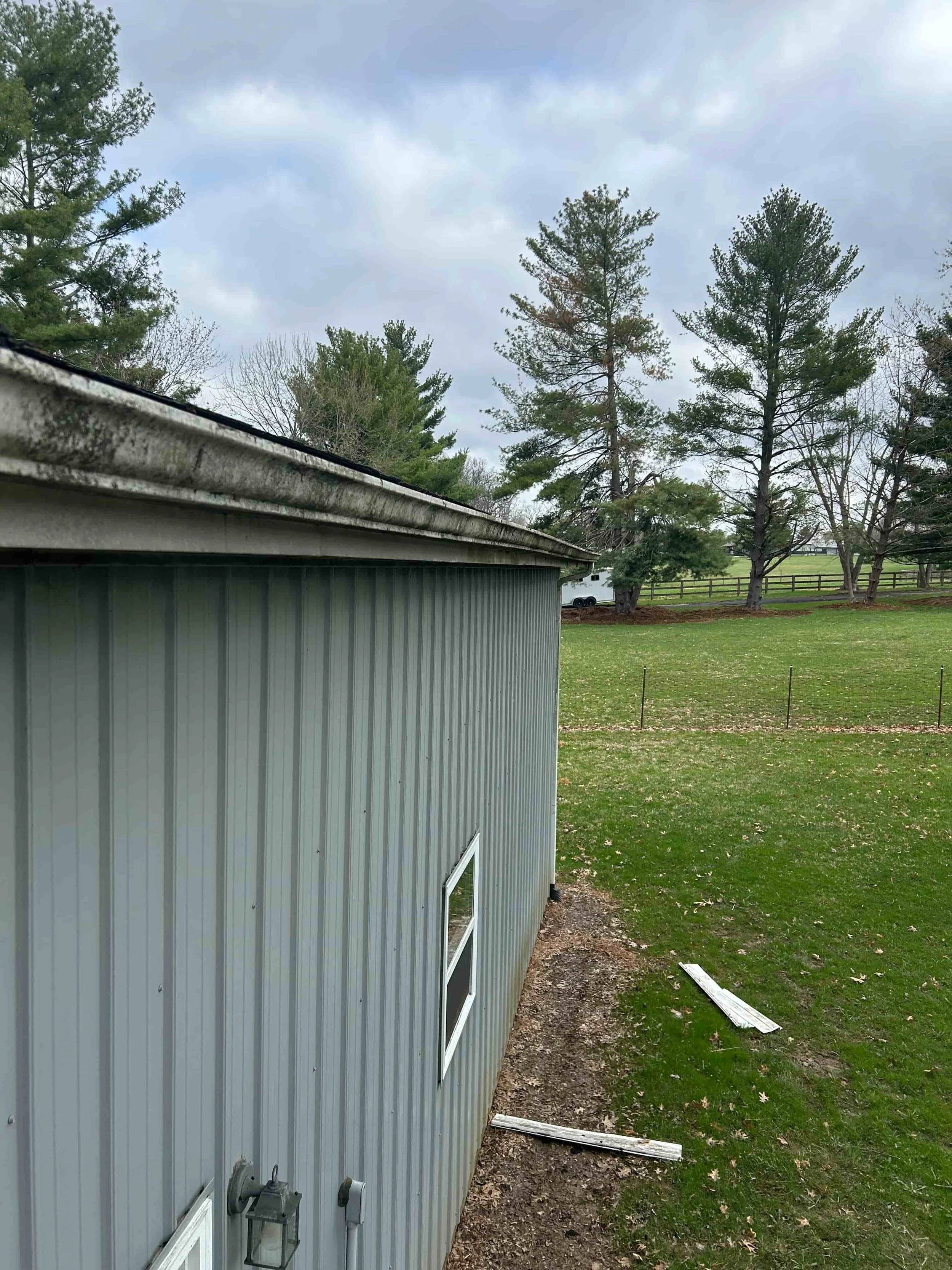 Side view of a brick house, showing a section of roof, gutter, and a gas meter. Green bushes line the house, and a lawn is visible in the background with trees and neighboring houses.