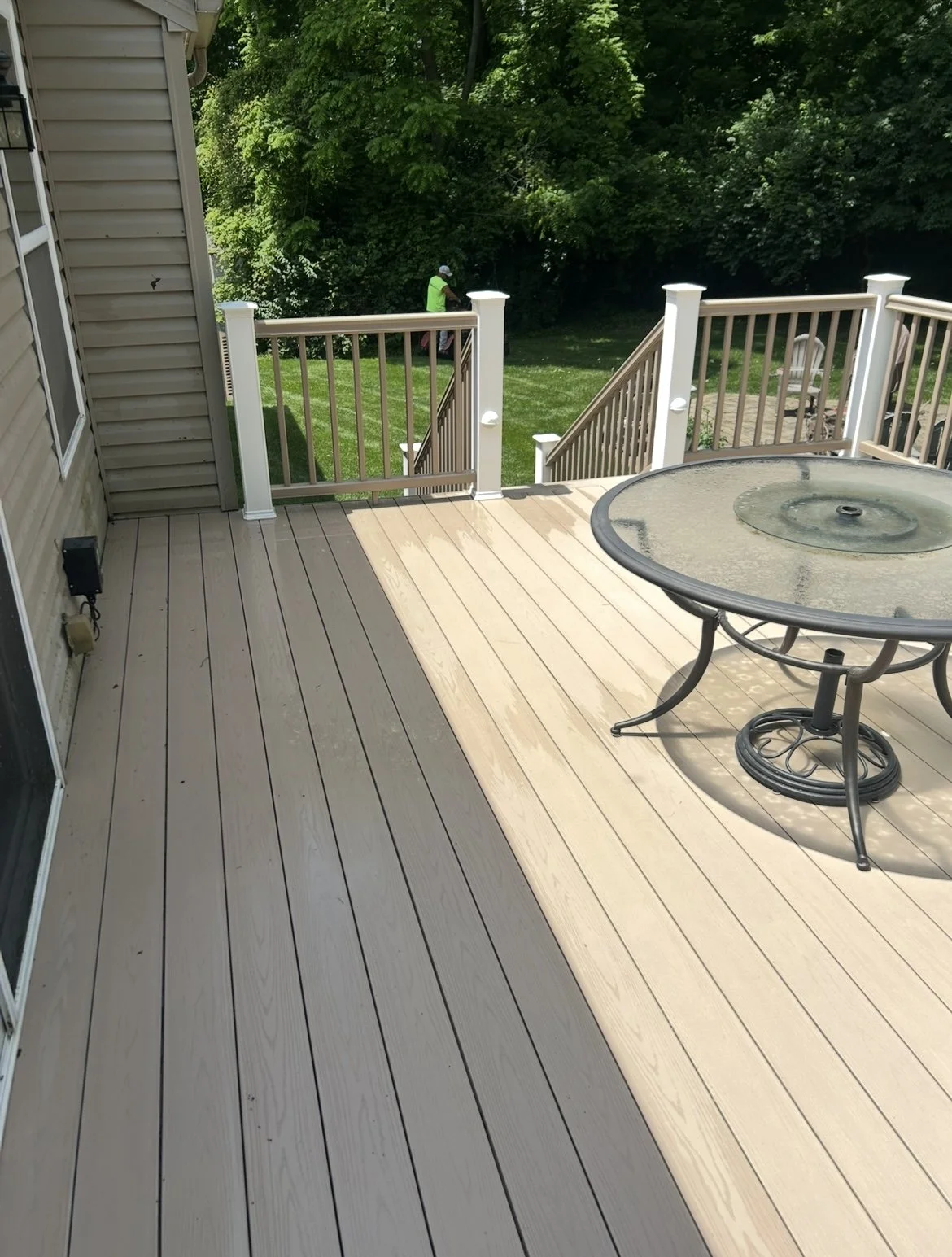 Back patio with a wooden deck, round glass table, and railing, overlooking a grassy lawn and trees, with a person in a neon vest and a cap working in the yard.