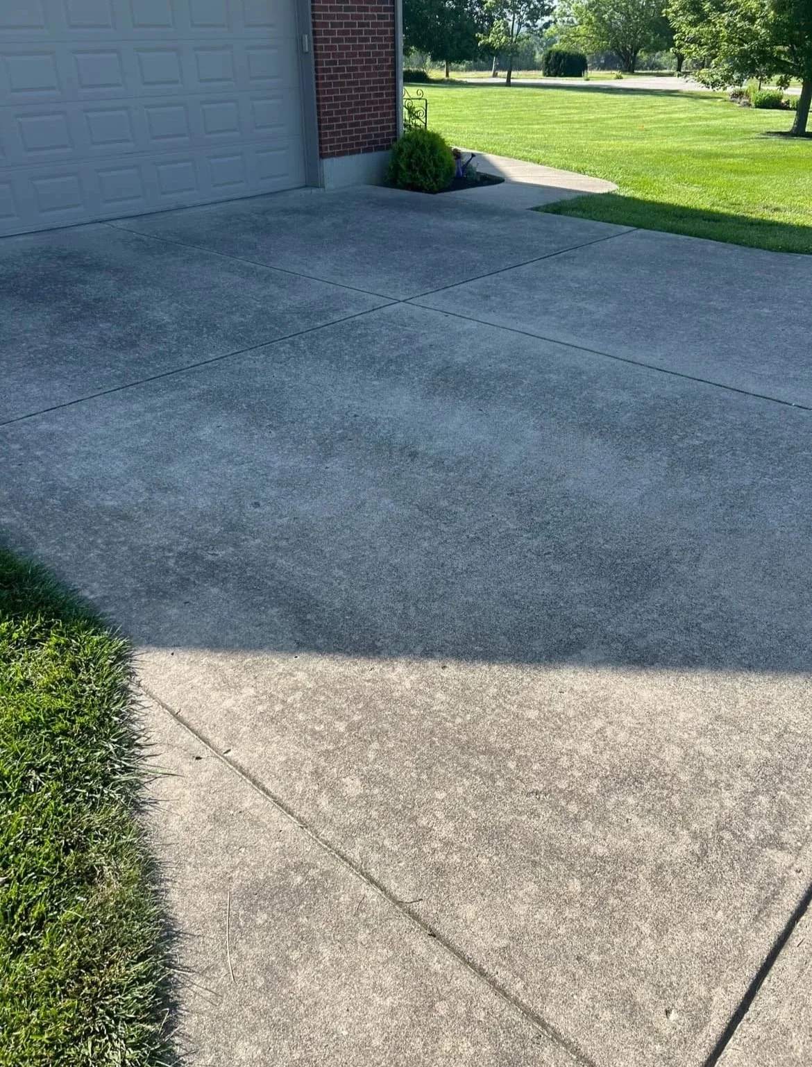 A residential driveway with grass on the left and right sides, with a garage door on the left and a small bush near the corner of the house, under a sunny sky.