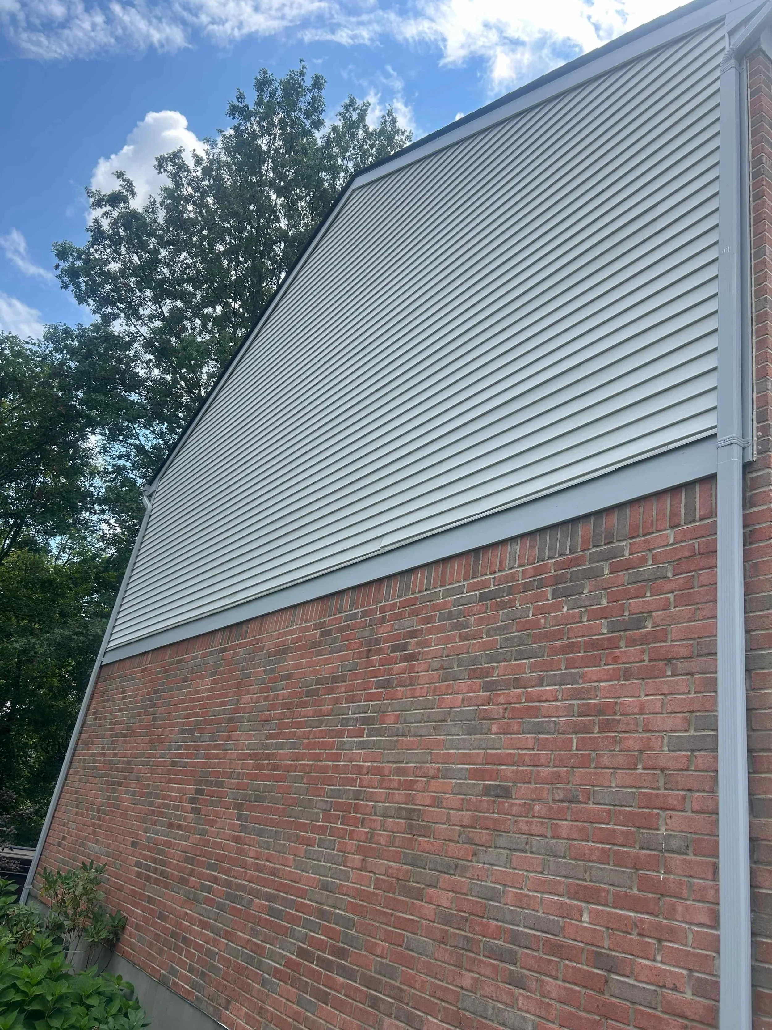 Close-up of a brick building with vinyl siding on the upper part, blue sky with scattered clouds, and trees in the background.