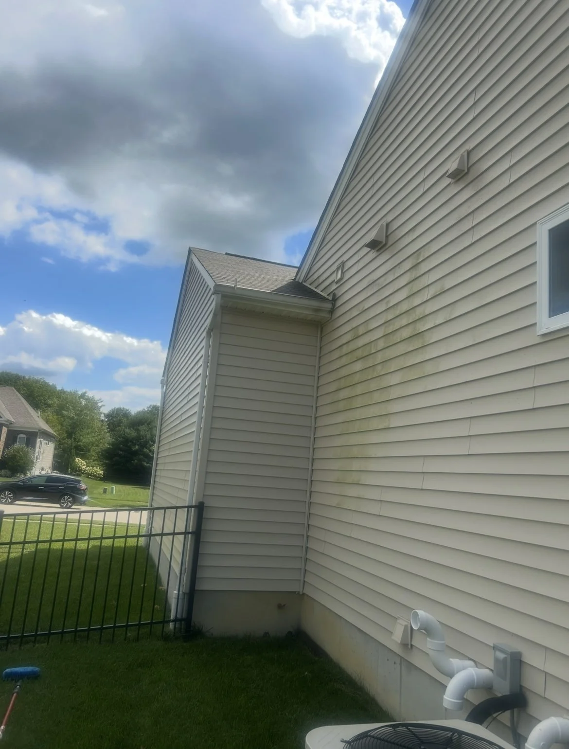 Side of a house with cream siding showing green mold or mildew on the wall, a vent, and a white PVC pipe in the yard, with a cloudy sky overhead.