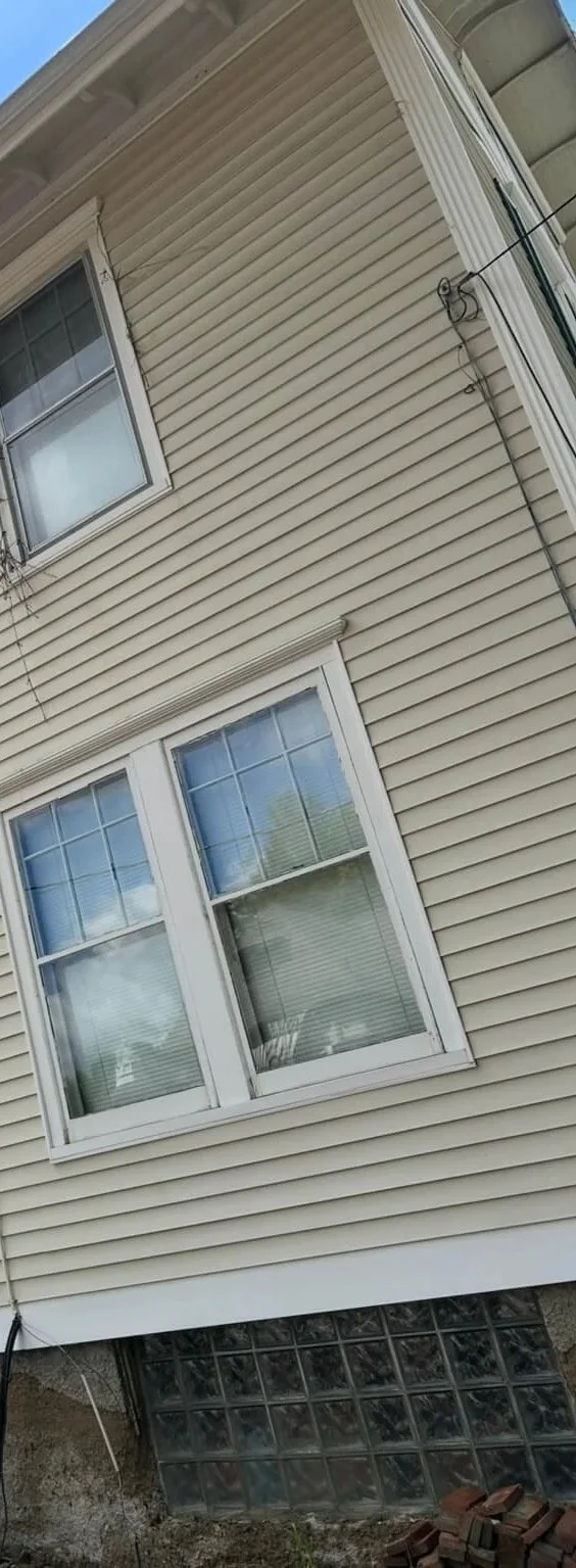 Close-up view of beige vinyl siding on a house with two white-framed windows, one above the other, and a basement window made of glass blocks at the bottom.