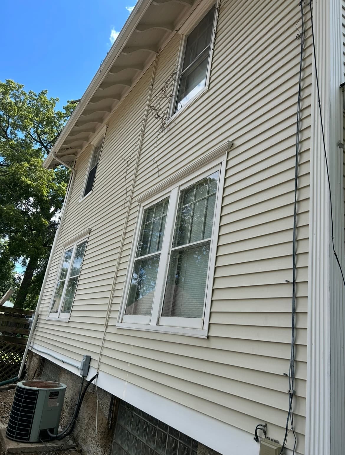Side view of a beige house with vinyl siding, several windows, and visible outdoor air conditioning unit. Blue sky and trees in the background.