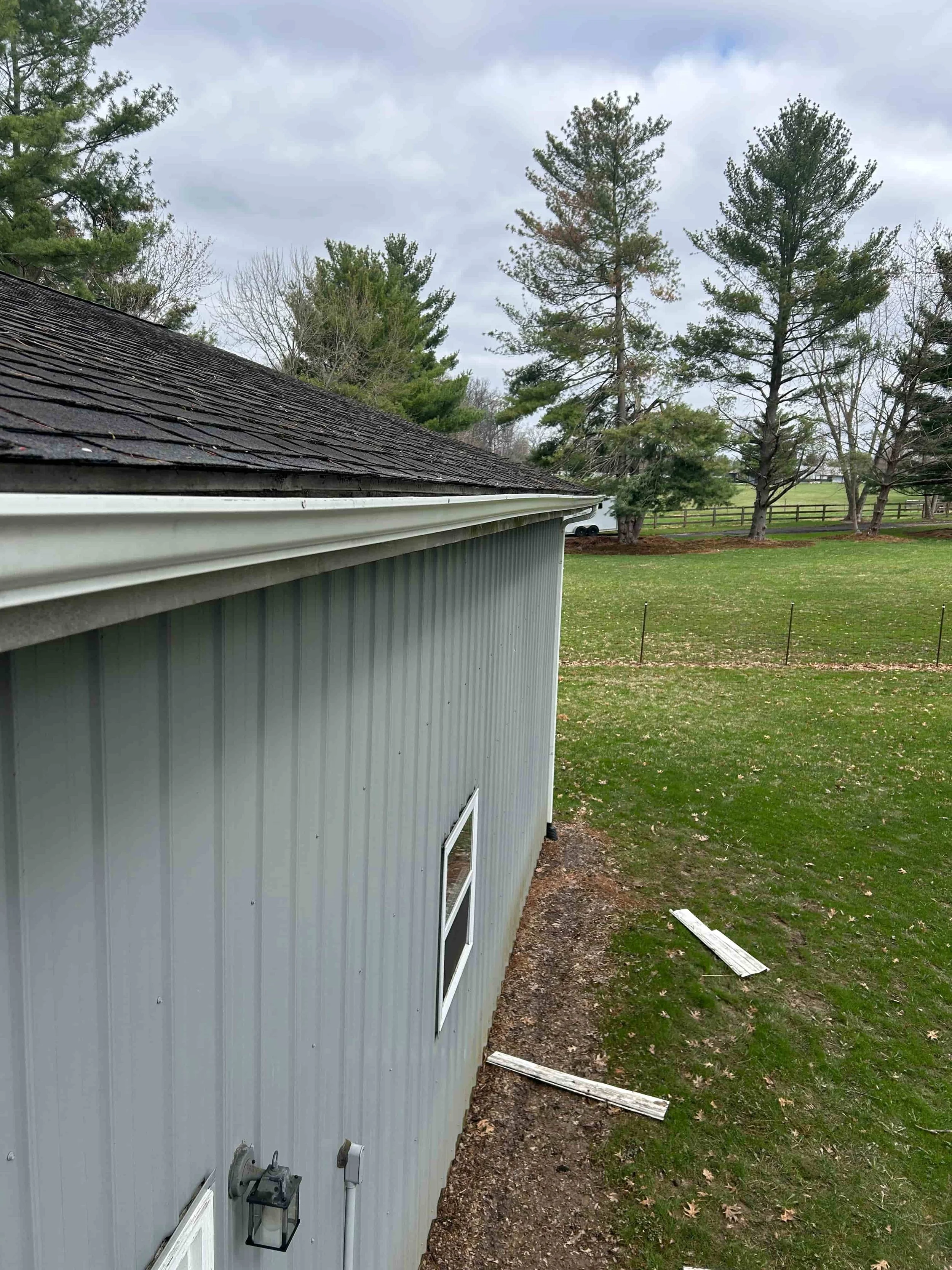 Side view of a gray metal building with a small window, situated on a grassy yard with trees and cloudy sky in the background.