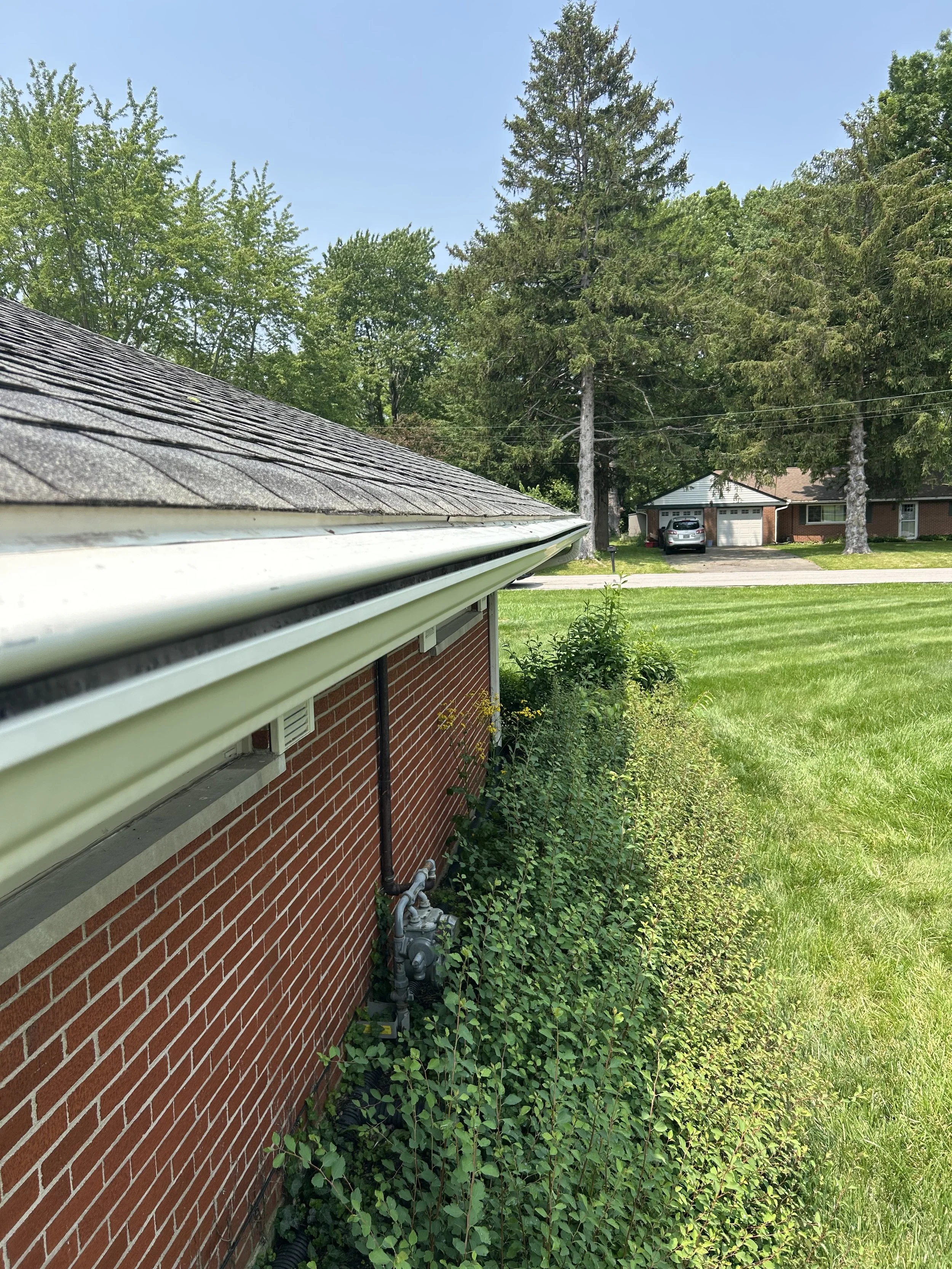 View of a suburban backyard with a brick house, green grass, and trees, with the roof and gutter of the house in the foreground and neighboring houses across the street.