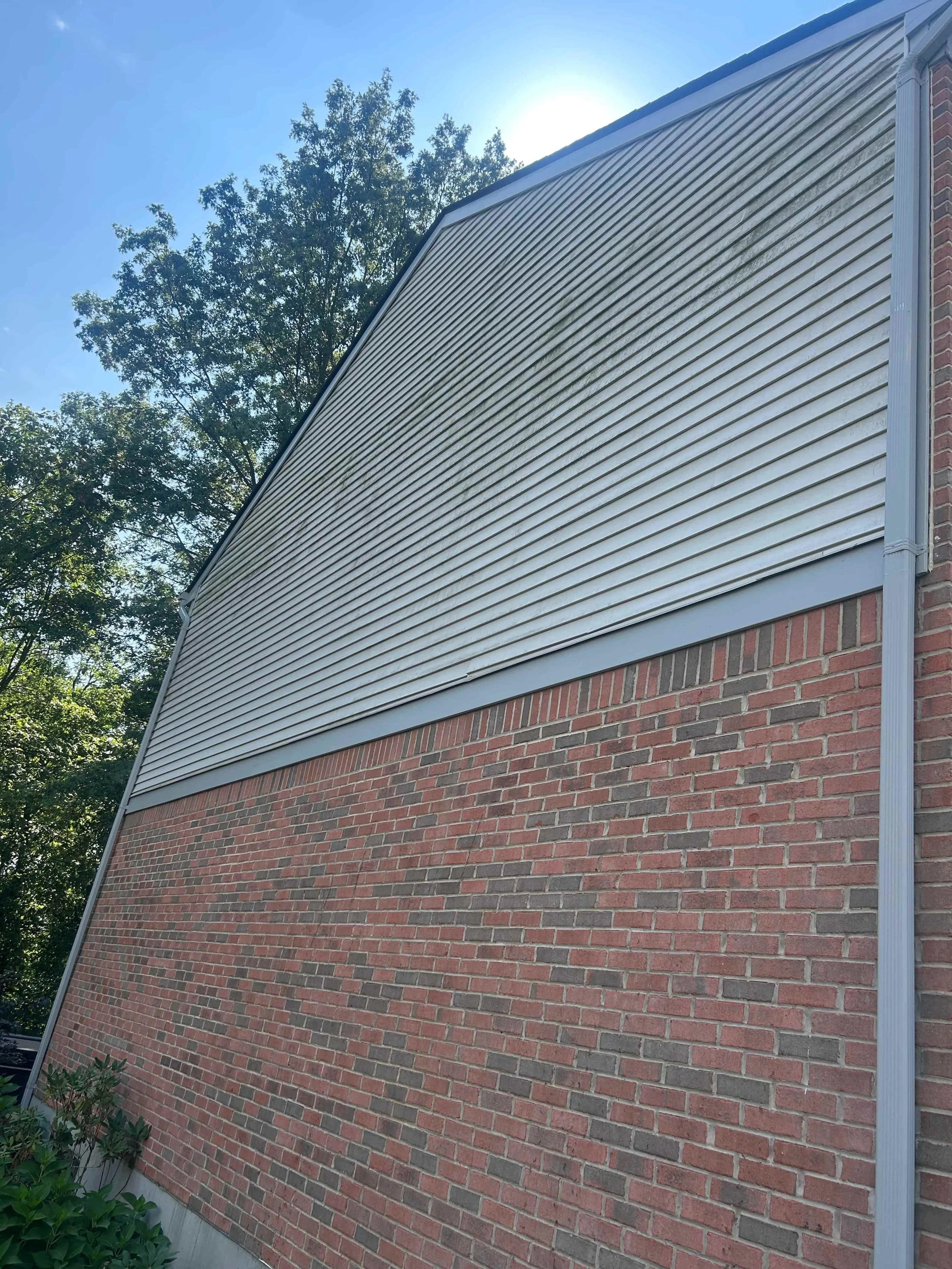 Close-up of a building wall showing red brick on the lower part and gray siding on the upper part, with a tree in the background and sunlight overhead.