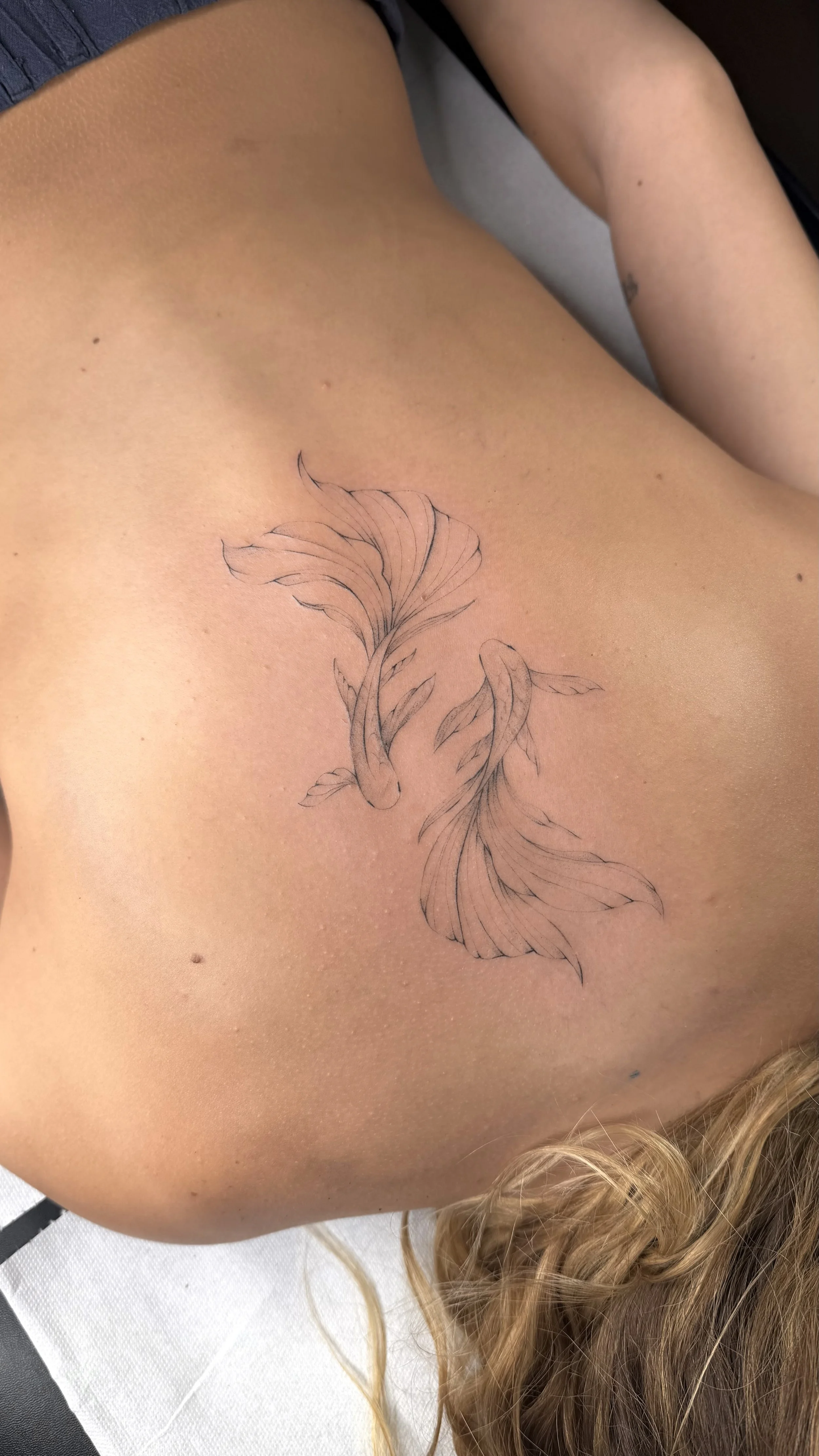 Tatouage de deux poissons en ligne fine sur la côte droite d'une personne aux cheveux bouclés et blondes, allongée sur une surface blanche