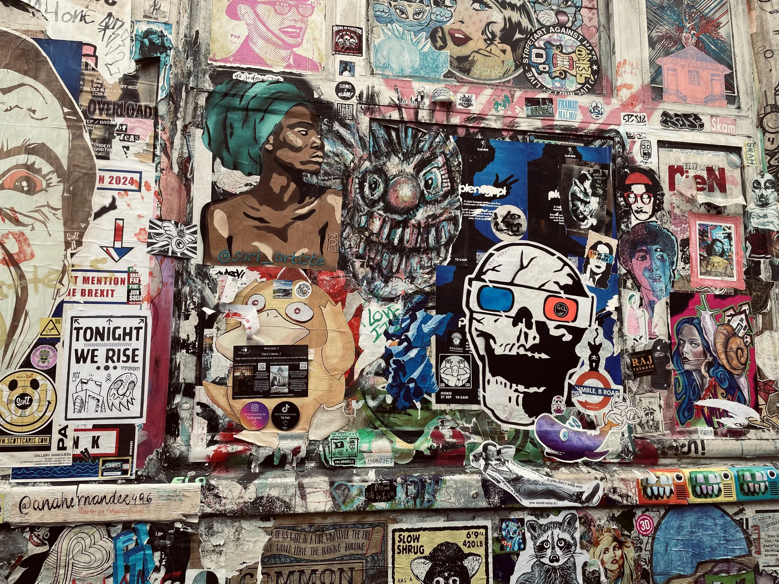 A wall covered with various colorful stickers, posters, and graffiti, including a prominent portrait of a woman with a headwrap, a large painted skull, and multiple smaller designs and text.