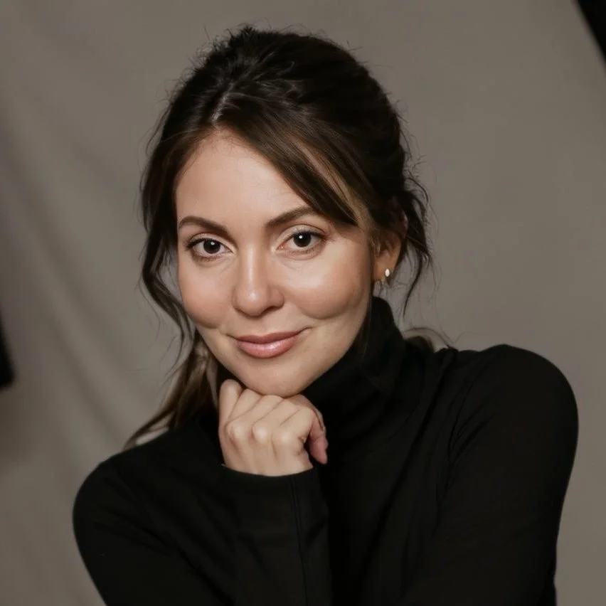 A woman with brown hair and fair skin, wearing a black turtleneck, posing with her chin resting on her hand and smiling gently at the camera.