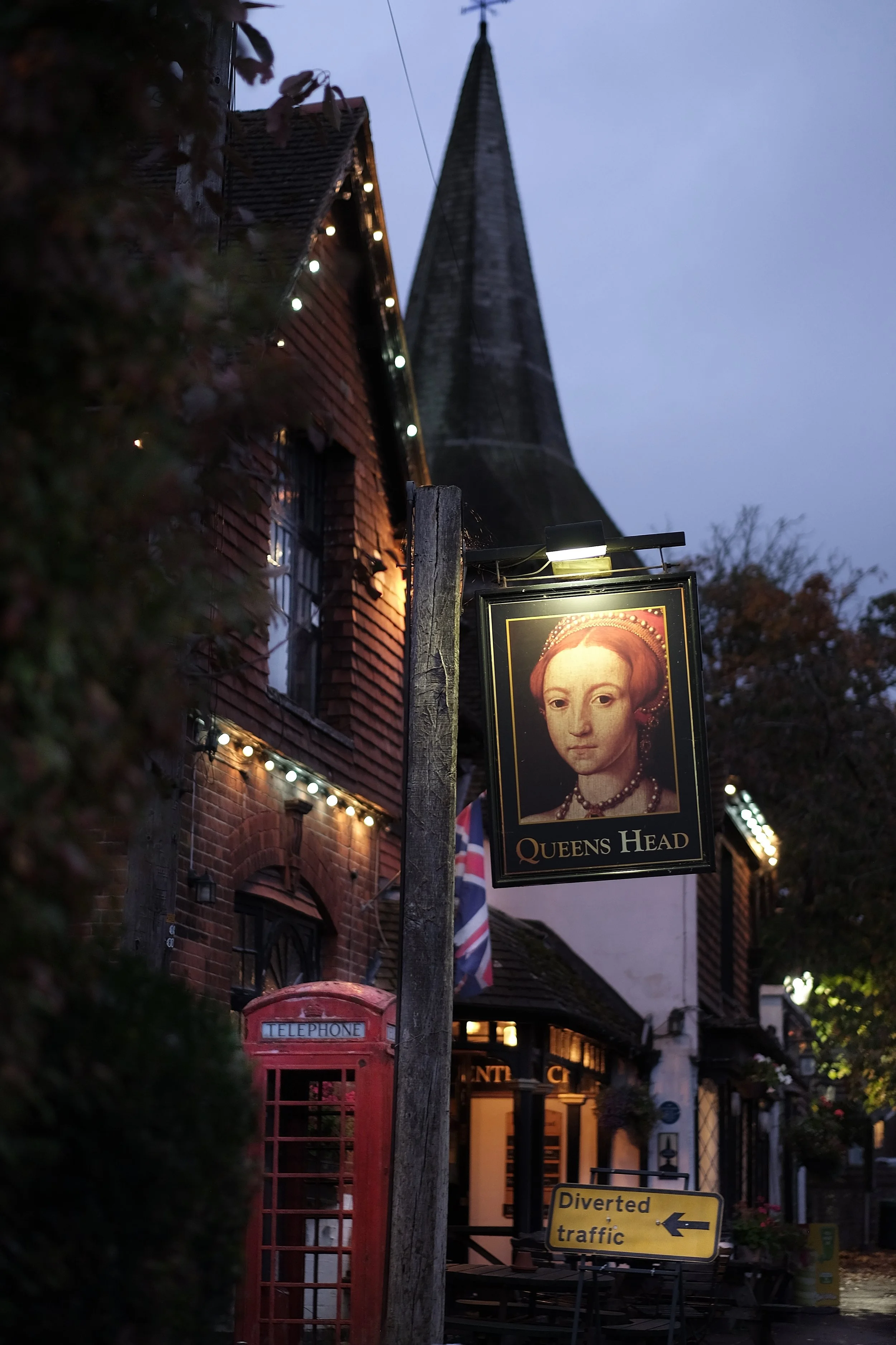 A street scene in a historic village with a large church steeple, hanging sign of Queen Elizabeth I, and a red British telephone booth, during evening hours with string lights illuminating the building.
