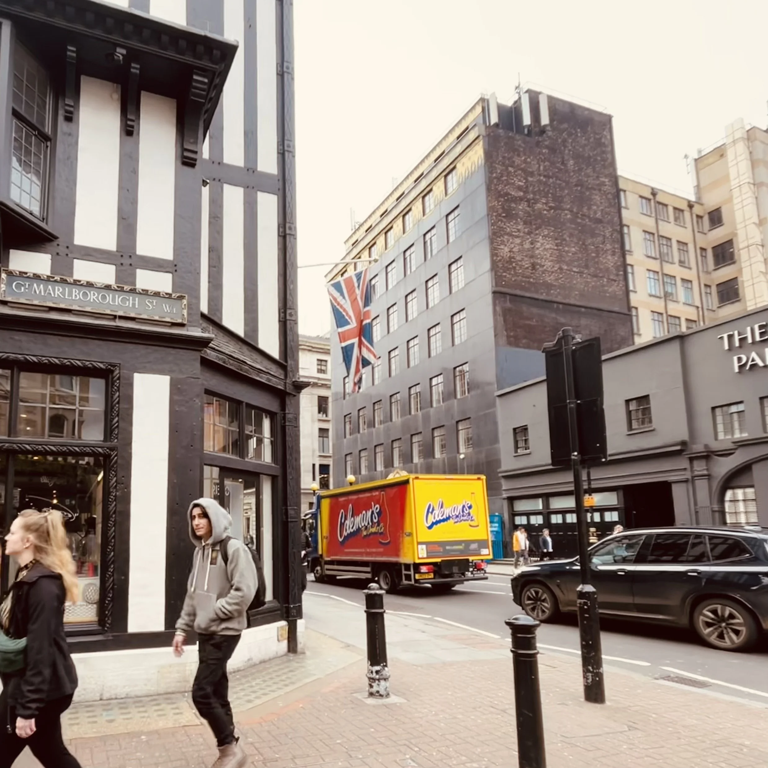 City street scene with pedestrians walking, parked cars, and buildings including a Union Jack flag and a street sign for G. Marlborough Street.