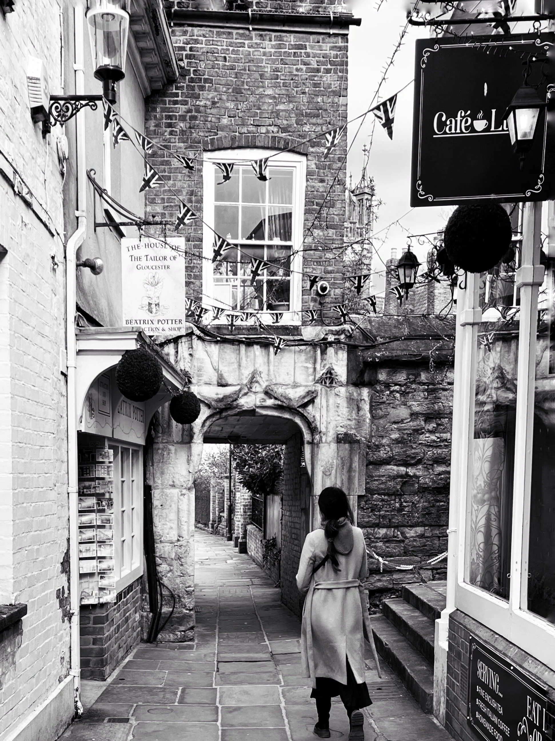 A narrow alleyway with brick and stone walls, decorated with Union Jack flags, leading to an archway. A woman with long dark hair, wearing a coat and boots, walks away from the camera. There are signs and shop windows on either side of the alley.