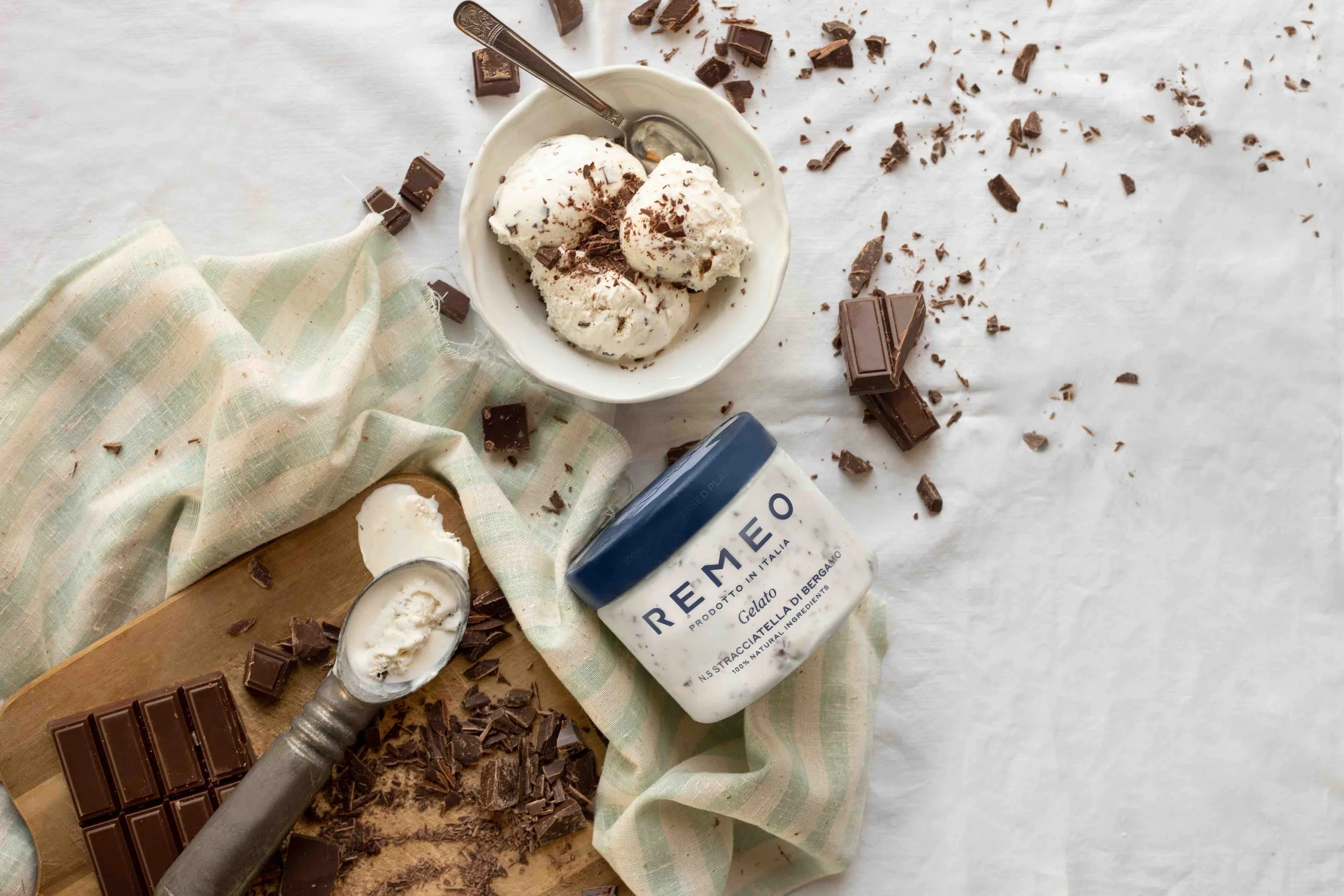 A bowl of vanilla ice cream with chocolate shavings, surrounded by broken chocolate bars, a jar of Reemoe gelato, a scoop of ice cream on a spoon, and a wooden cutting board, all on a white surface with a checkered cloth.