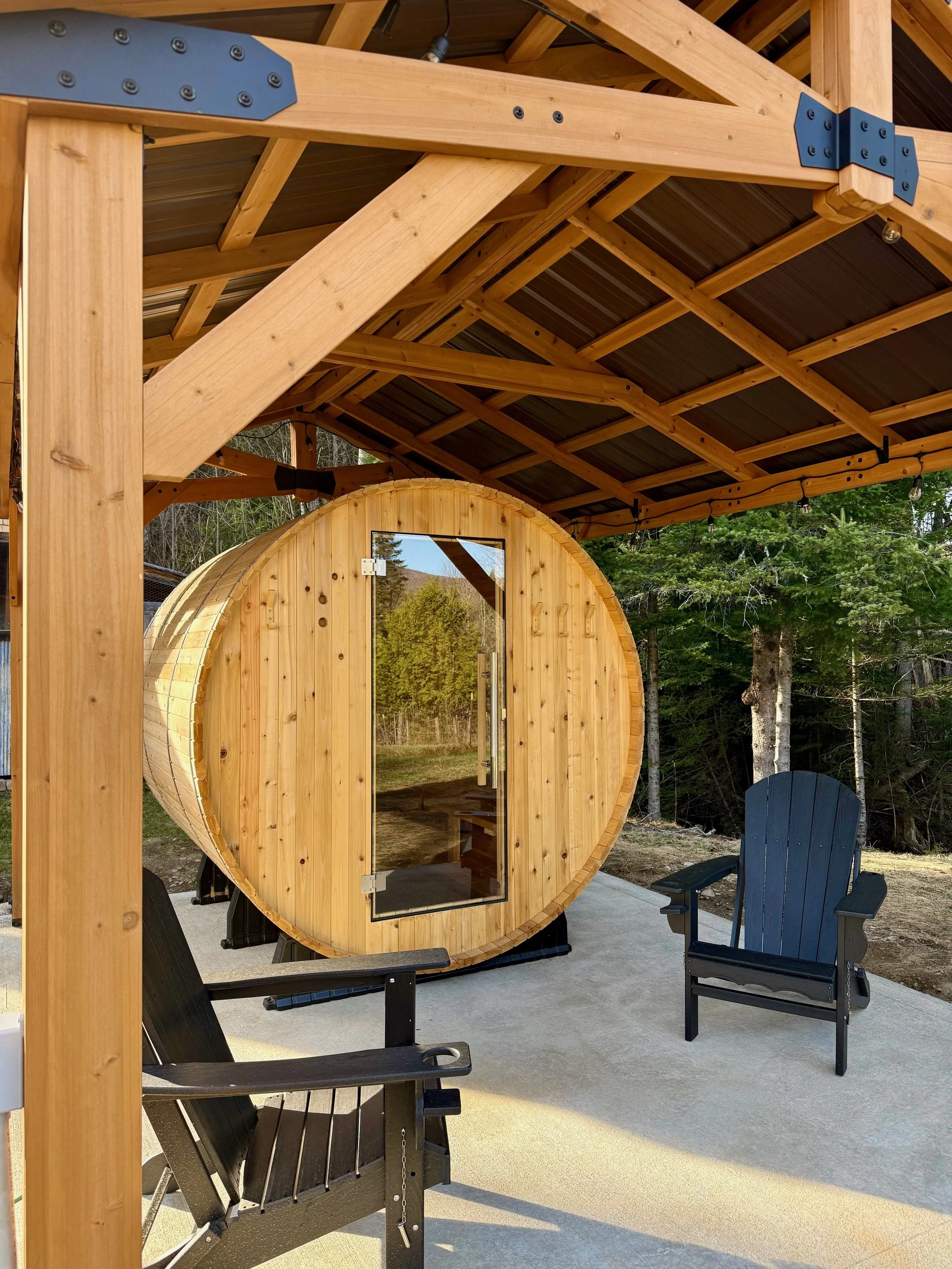 A wooden barrel-shaped sauna with a glass door set on snow outside, surrounded by snow-covered trees and two black Adirondack chairs nearby.