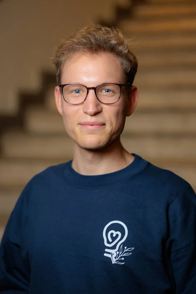 Person wearing glasses and a blue sweatshirt with a heart and leaf logo, standing in front of blurry steps.