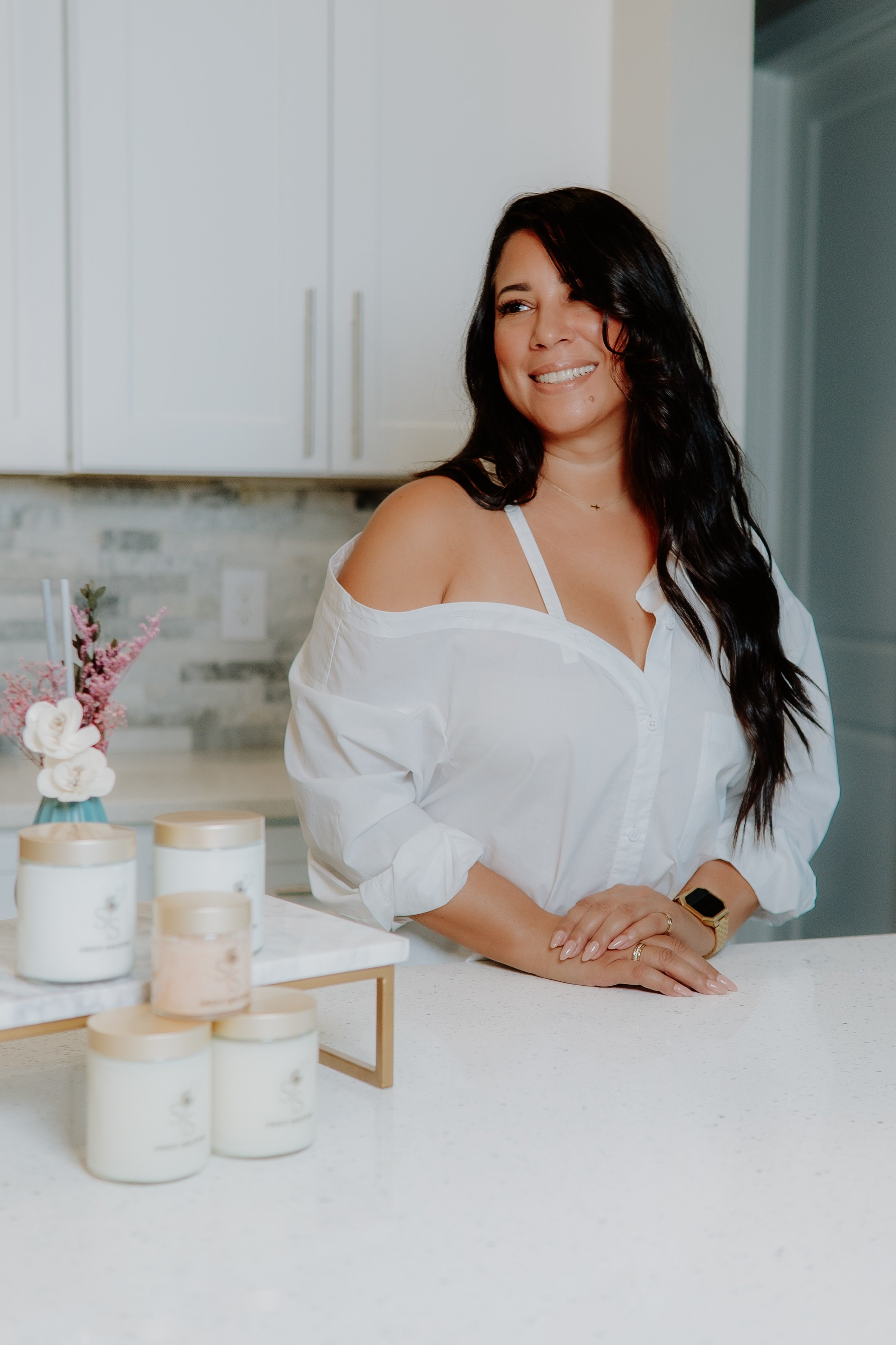 A woman with long dark hair smiling in a white blouse, standing at a kitchen island with candles and a flower arrangement.