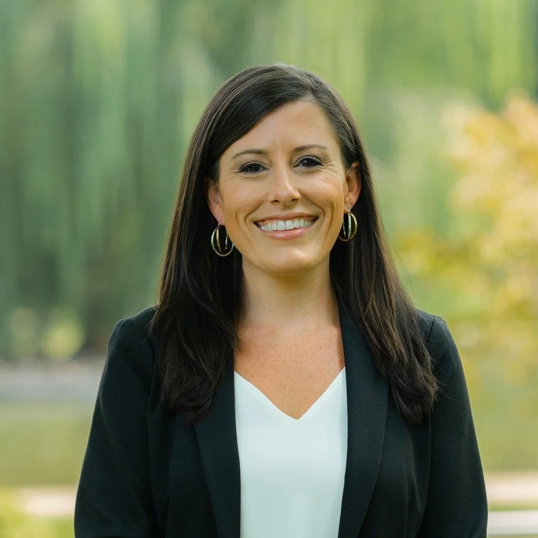 Portrait of a smiling woman with dark brown hair, wearing hoop earrings, a black blazer, and a white top, outdoors with blurred green trees in the background.