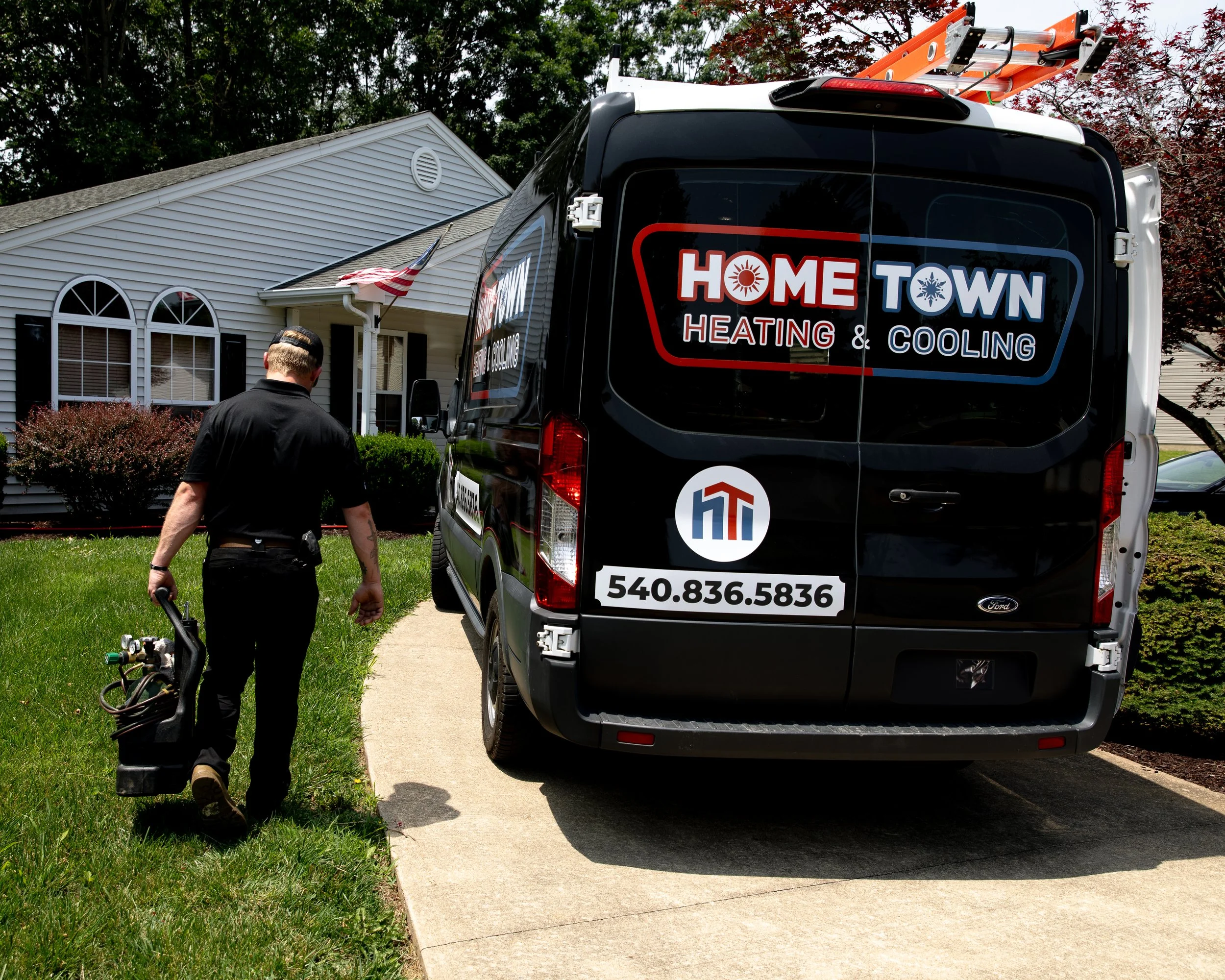 A technician walking on a lawn next to a driveway, carrying equipment, with a service van labeled 'Home Town Heating & Cooling' parked nearby in front of a house.