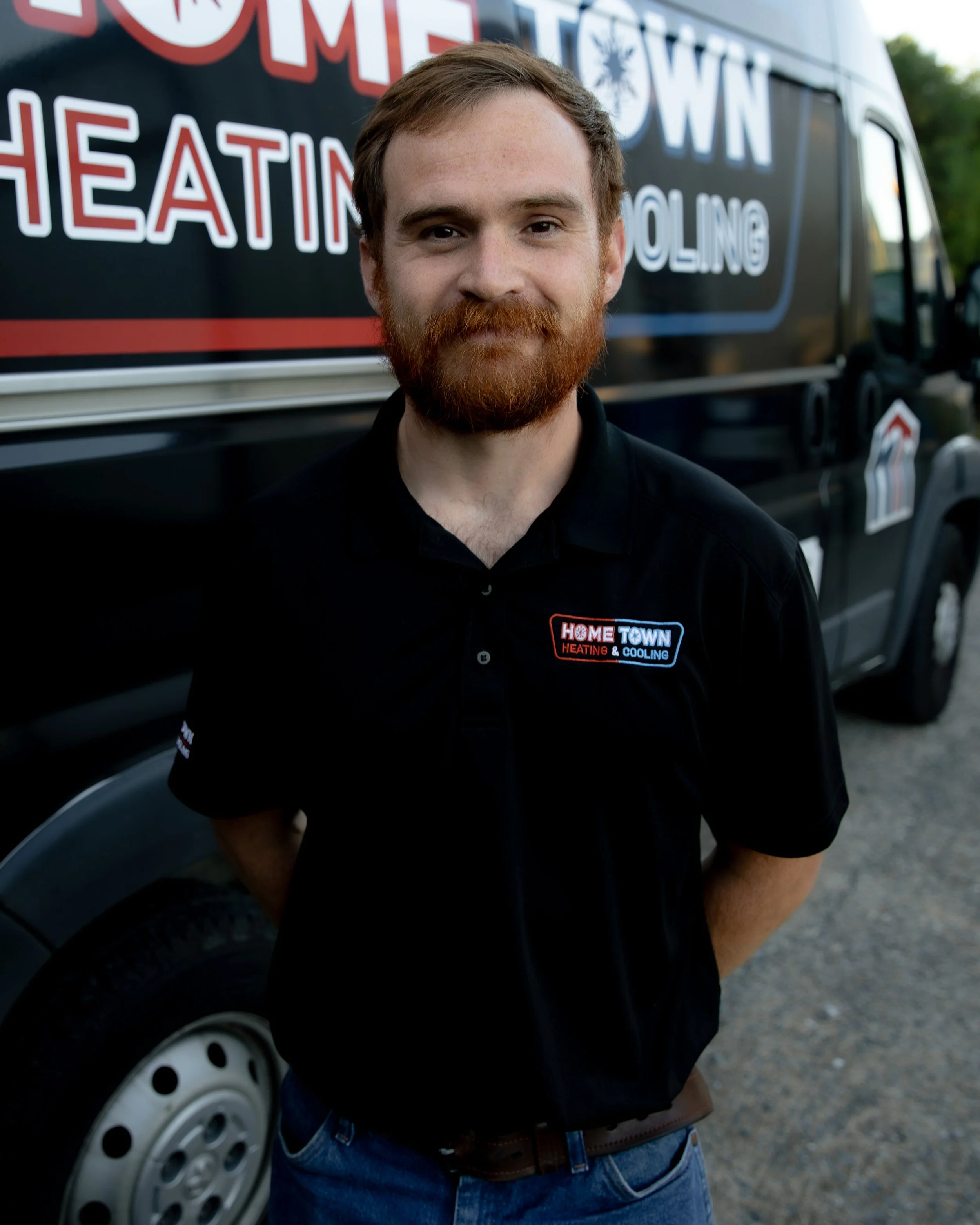 A man with red hair and beard smiling, standing outdoors in front of a black service van with 'Home Town Heating & Cooling' signage.