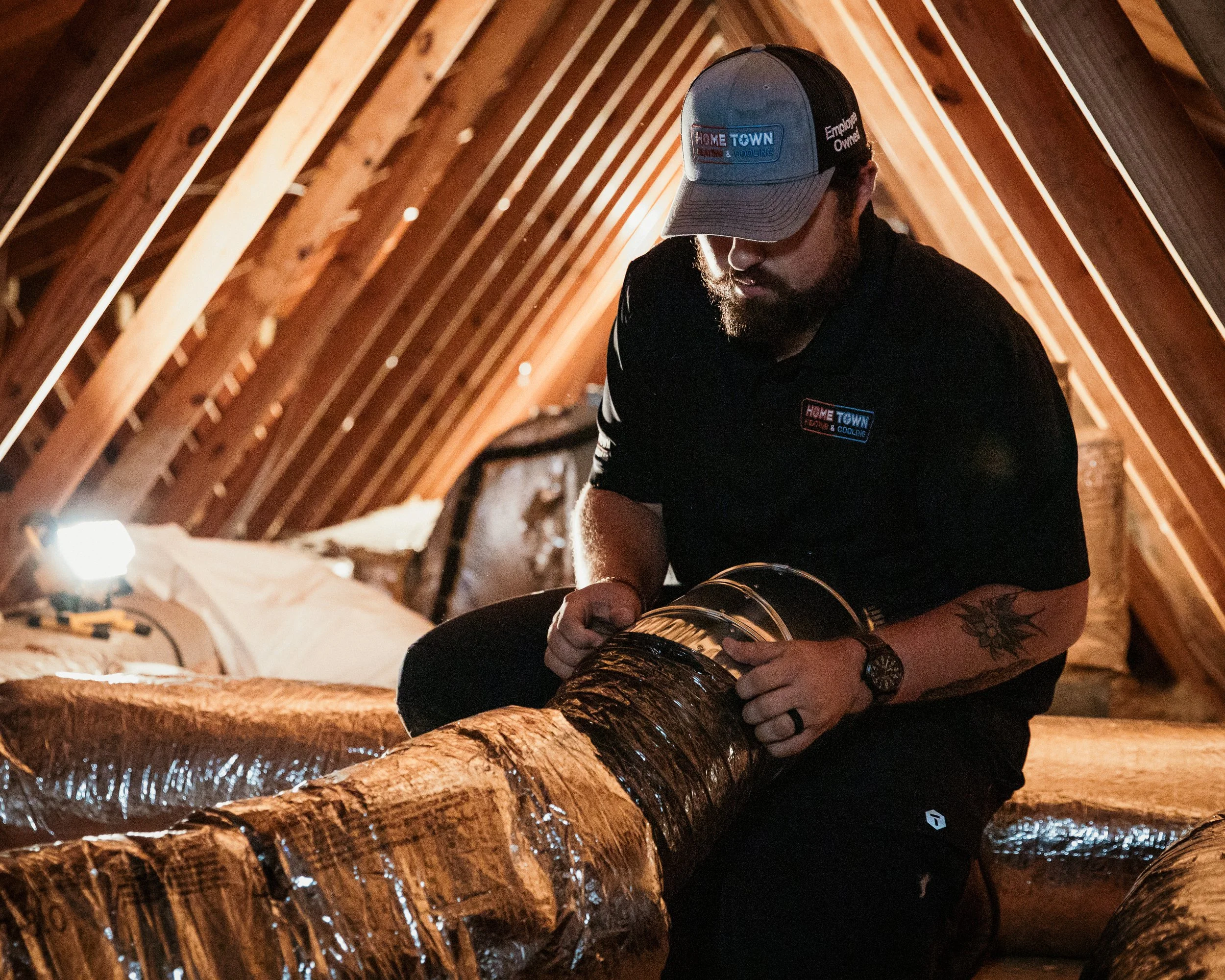 A man working on HVAC duct insulation inside an attic. He is wearing a black shirt and a baseball cap with company logos, holding a section of insulated duct.